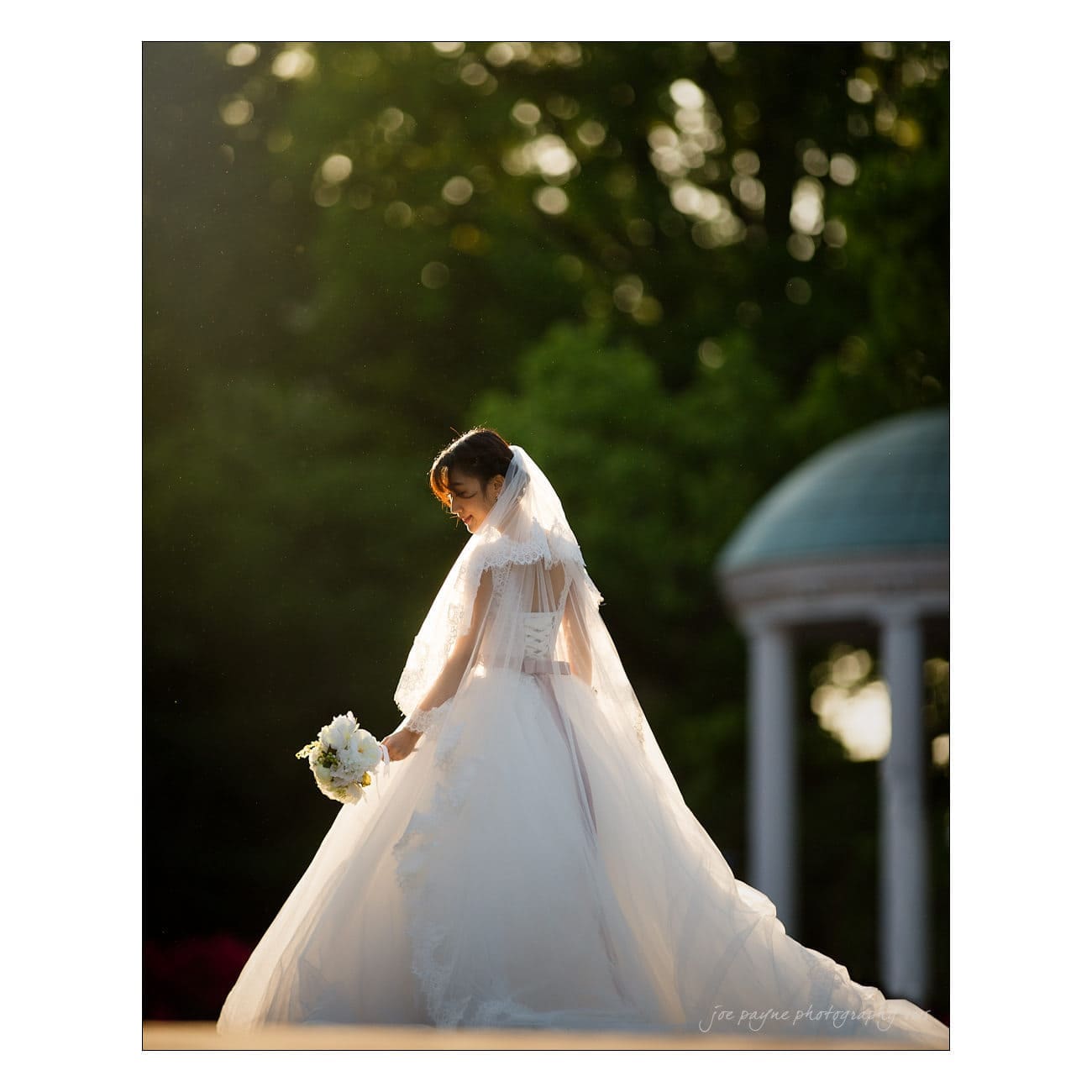 A bride in a flowing white dress and veil holds a bouquet while standing outdoors. Sunlight filters through the trees, creating a soft glow around her. A gazebo is visible in the background.