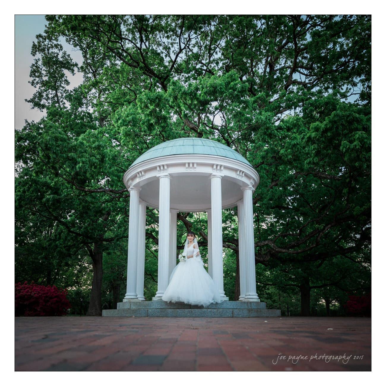 A bride in a white gown stands in the center of a classical white gazebo with columns and a domed roof, surrounded by lush green trees. The brick ground contrasts with the vibrant foliage.