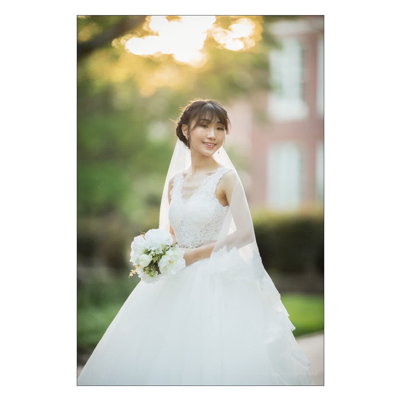 A bride in a white wedding dress smiles outdoors. She holds a bouquet of white flowers, and her veil cascades behind her. The background is softly blurred with greenery and sunlit trees.