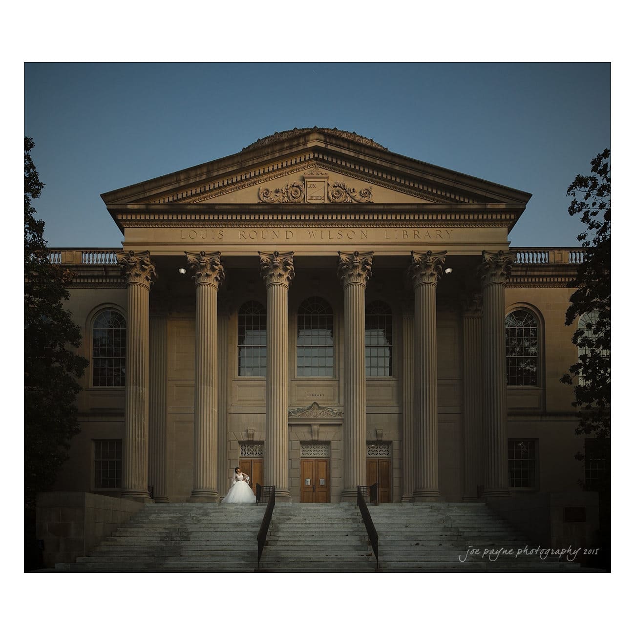 A bride and groom stand on the steps of the Louis Round Wilson Library, an impressive neoclassical building with six large columns and a pediment. The evening sky adds a serene backdrop to the scene.