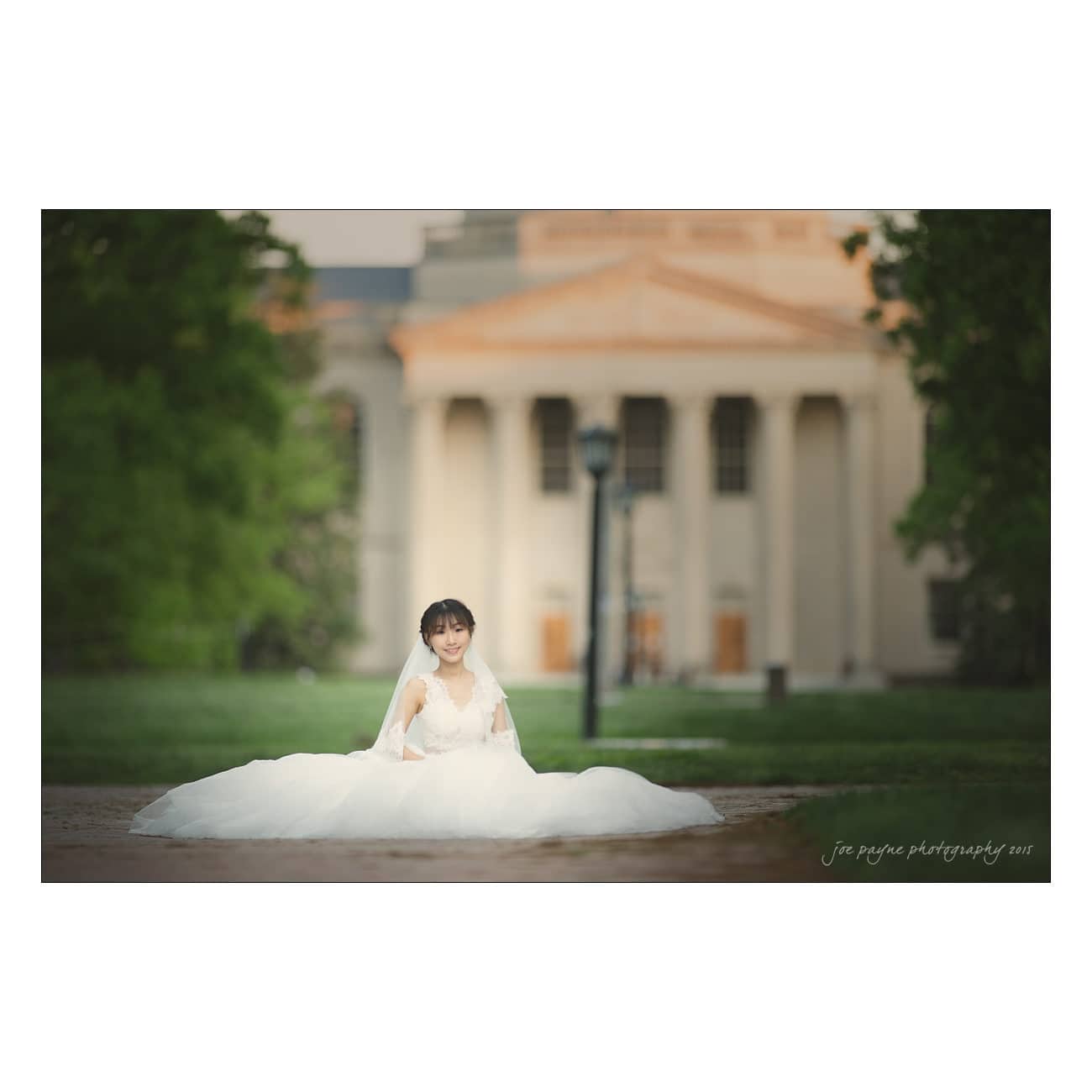 A bride in a flowing white gown sits on a path in front of a grand building with columns. The background is lush with greenery, suggesting a peaceful and elegant setting. Her veil drapes gracefully over her shoulders.