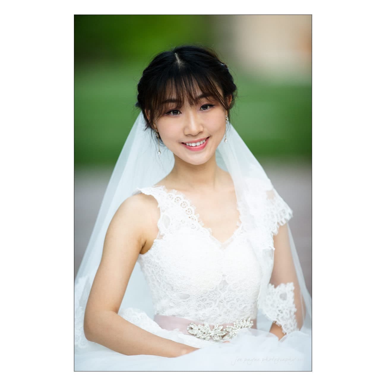 A smiling bride in a white lace wedding dress and veil poses outdoors. Her hair is styled in an elegant updo, and she wears delicate earrings. The background is softly blurred with hints of green.