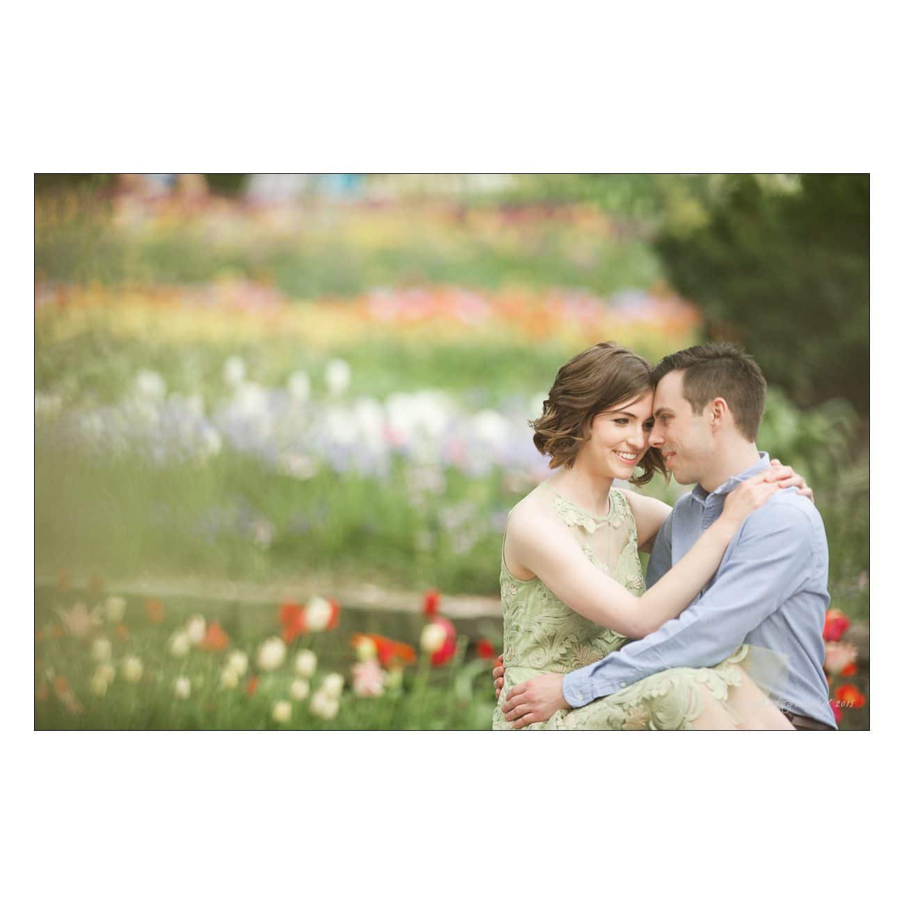 A couple sits closely in a garden filled with colorful flowers. The woman, in a green lace dress, gently leans on the mans shoulder. The scene conveys a sense of tenderness and romance.