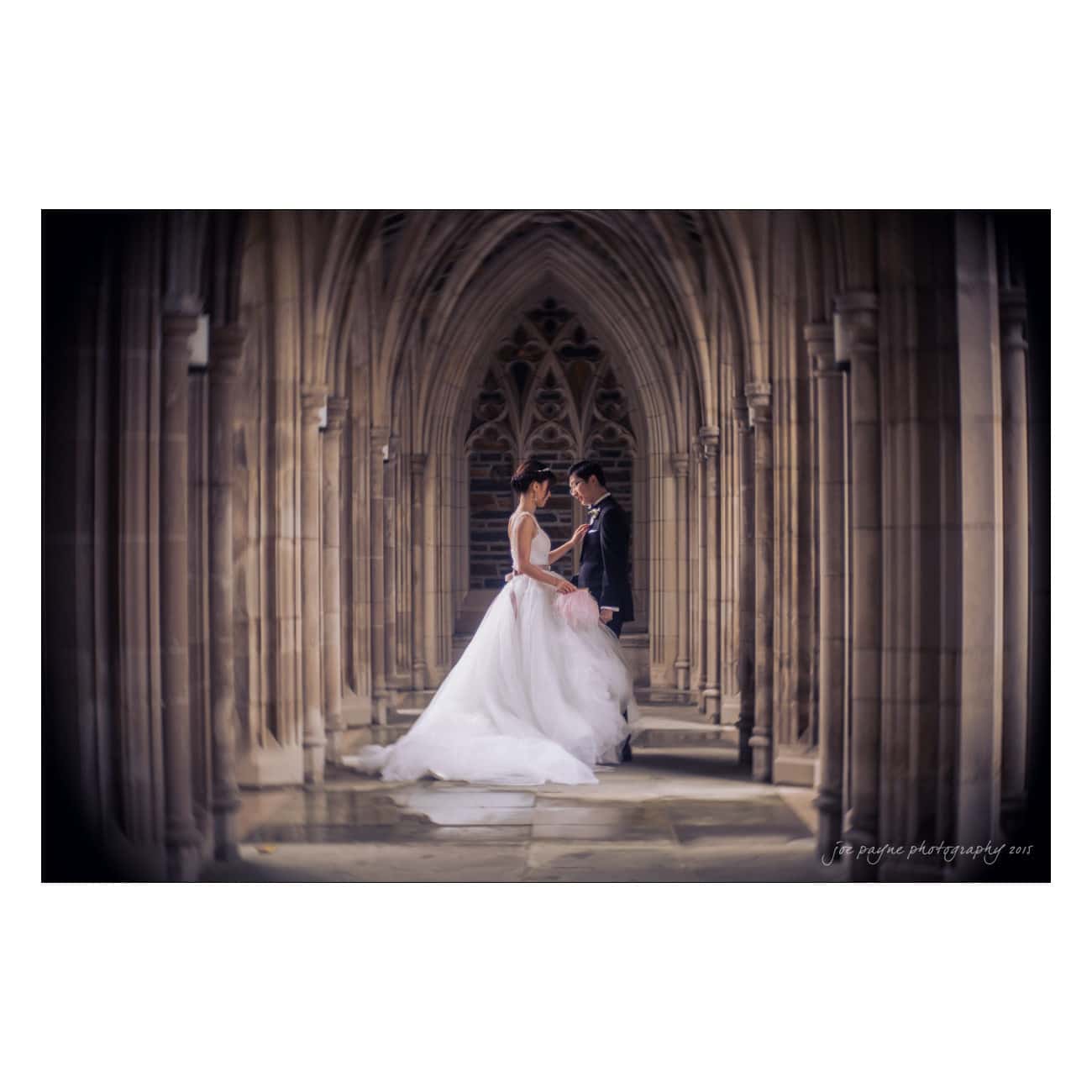 A bride and groom stand facing each other in a Gothic-style stone corridor, capturing a romantic moment. The bride wears a flowing white gown, and the groom is in a dark suit. The arches create a dramatic and intimate backdrop.