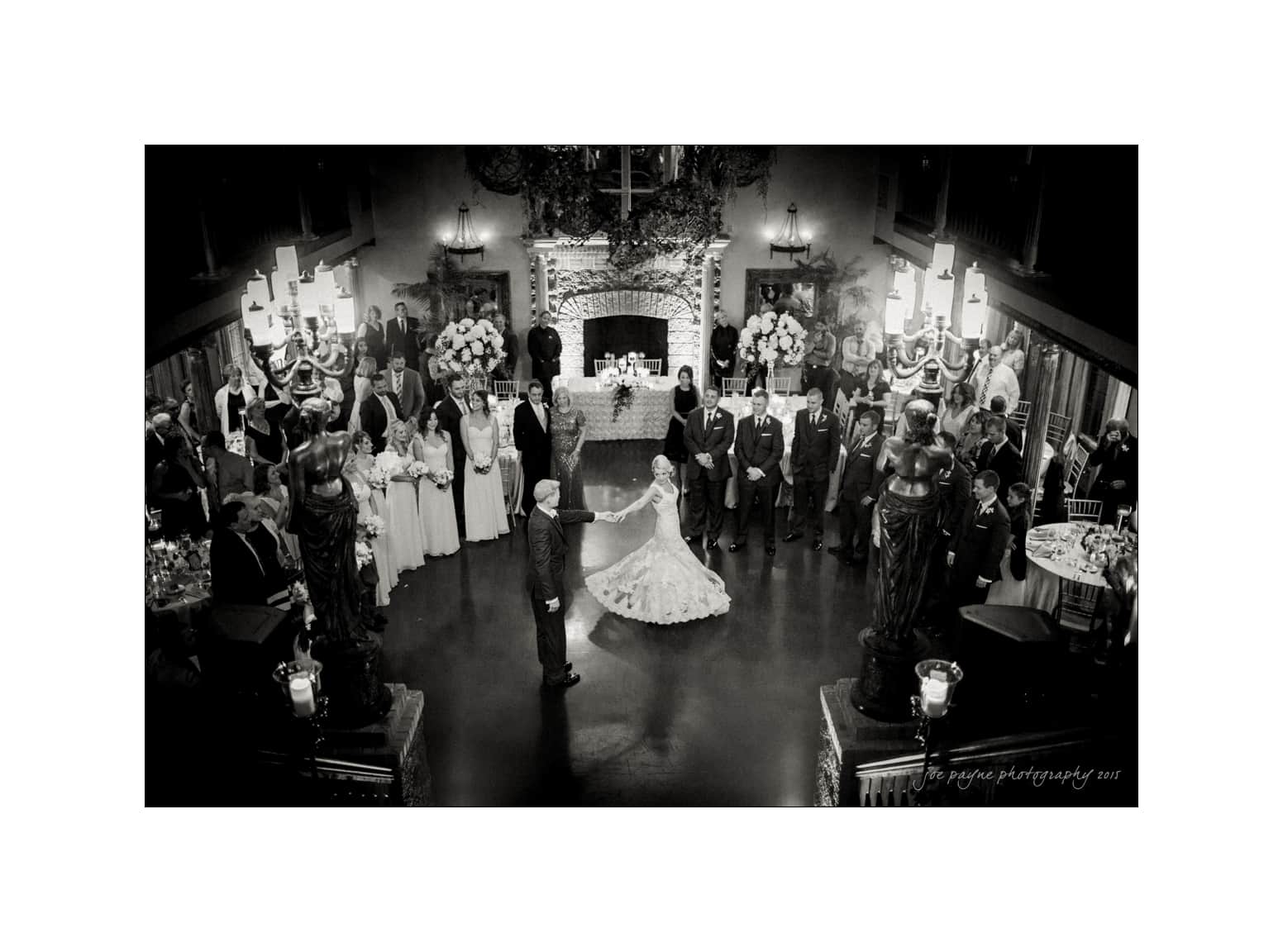 A black-and-white image of a wedding reception with a bride and groom dancing in the center of a large room. Guests surround them, watching. The room is elegantly decorated, with floral arrangements and ornate lighting.