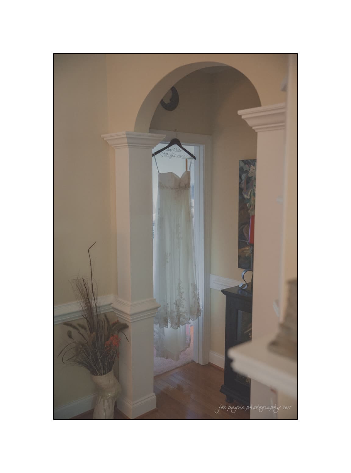 NC wedding photography - rachel & joe A white wedding dress hangs on a wooden hanger in an arched doorway. The dress is adorned with lace details and is in a softly lit room with a vase of dried flowers and a piece of colorful artwork on the wall.