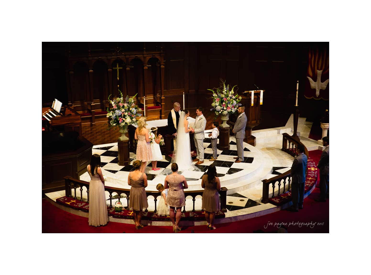 NC wedding photography - rachel & joe A wedding ceremony with a couple exchanging vows on a circular platform. They are surrounded by a flower-decorated arch, a pastor, and several attendants. Guests are watching from the aisles in a dimly lit chapel.