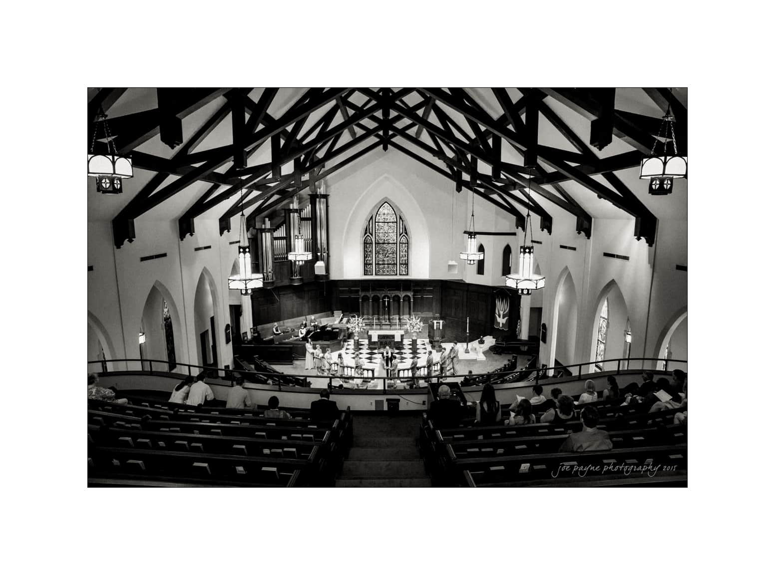 NC wedding photography - rachel & joe Black and white photo of a large church interior, featuring wooden beams and arched windows. The view is from the balcony, overlooking empty pews and a stage with a piano and music stands. Soft lighting highlights the architectural details.