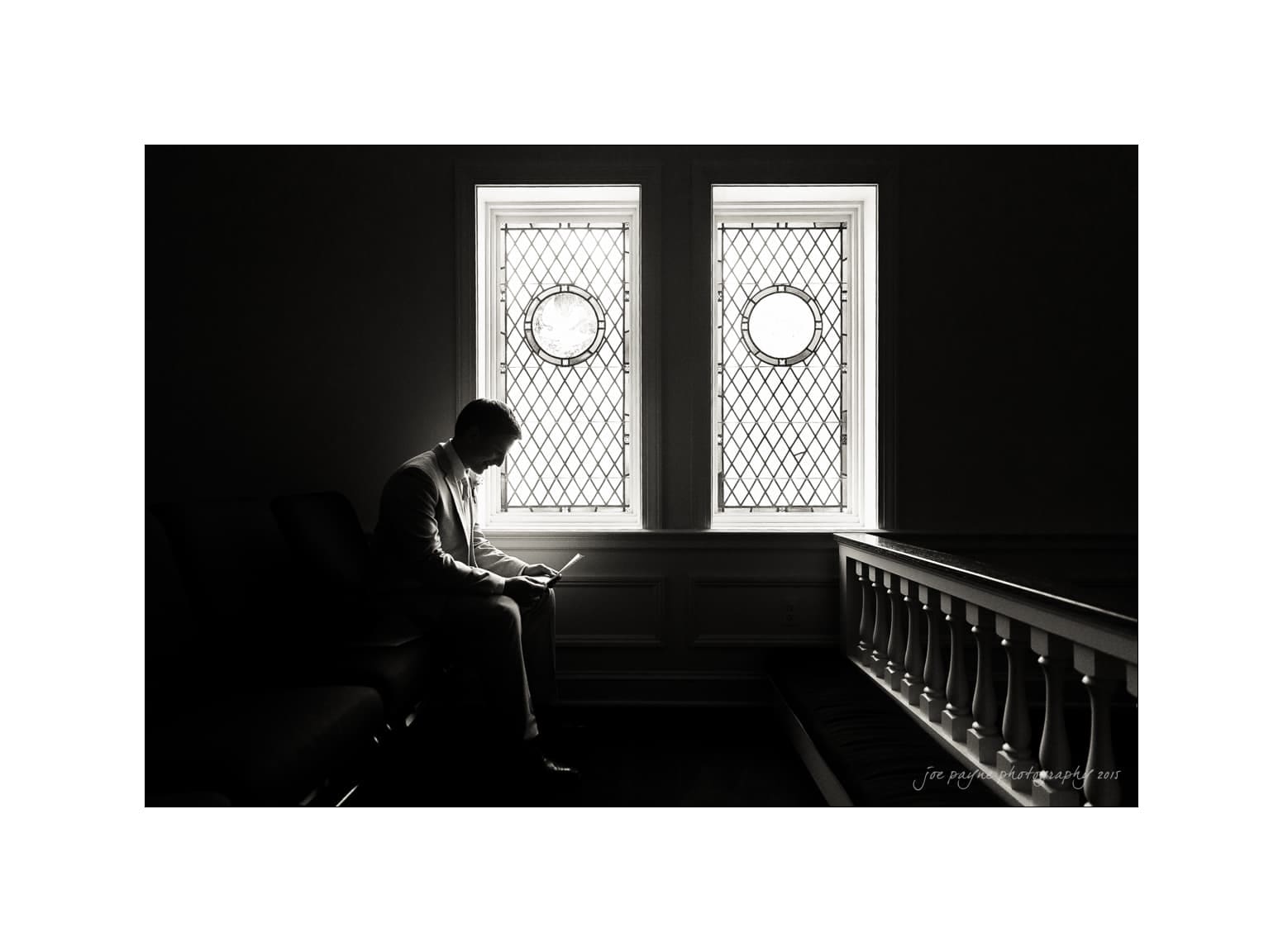 NC wedding photography - rachel & joe A man sits in a dimly lit room, silhouetted against two ornate stained glass windows, reading a book on a bench. The light from the windows creates a dramatic contrast with the shadowy surroundings.