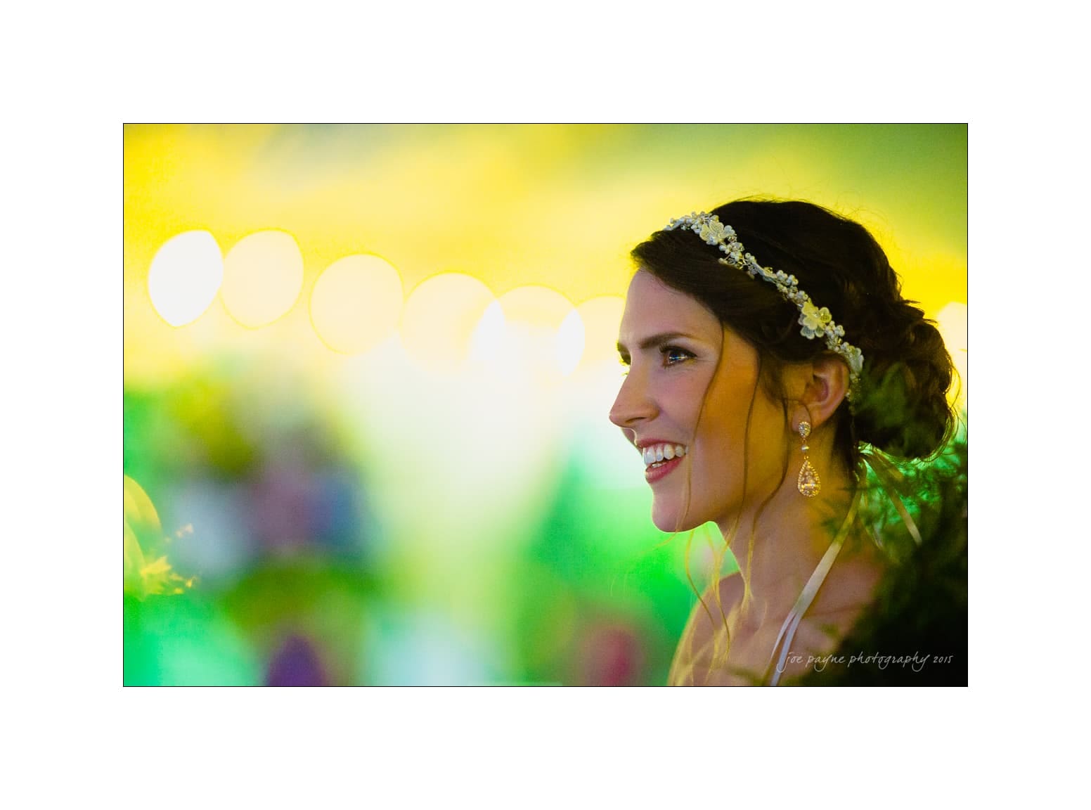 NC wedding photography - rachel & joe A smiling bride with a floral hairpiece and drop earrings, captured in profile against a softly blurred, colorful background with glowing lights.