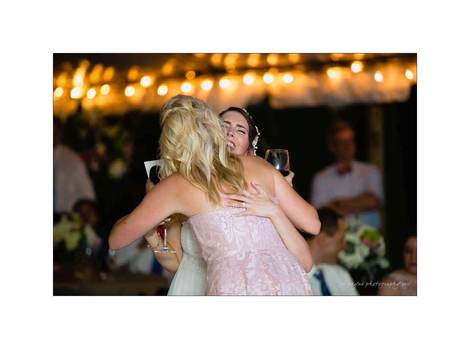 NC wedding photography - rachel & joe Two women wearing elegant dresses embrace at a festive event. The background is softly lit with string lights, and guests are visible in the blurred background, creating a warm and joyful atmosphere. One woman holds a wine glass in her hand.