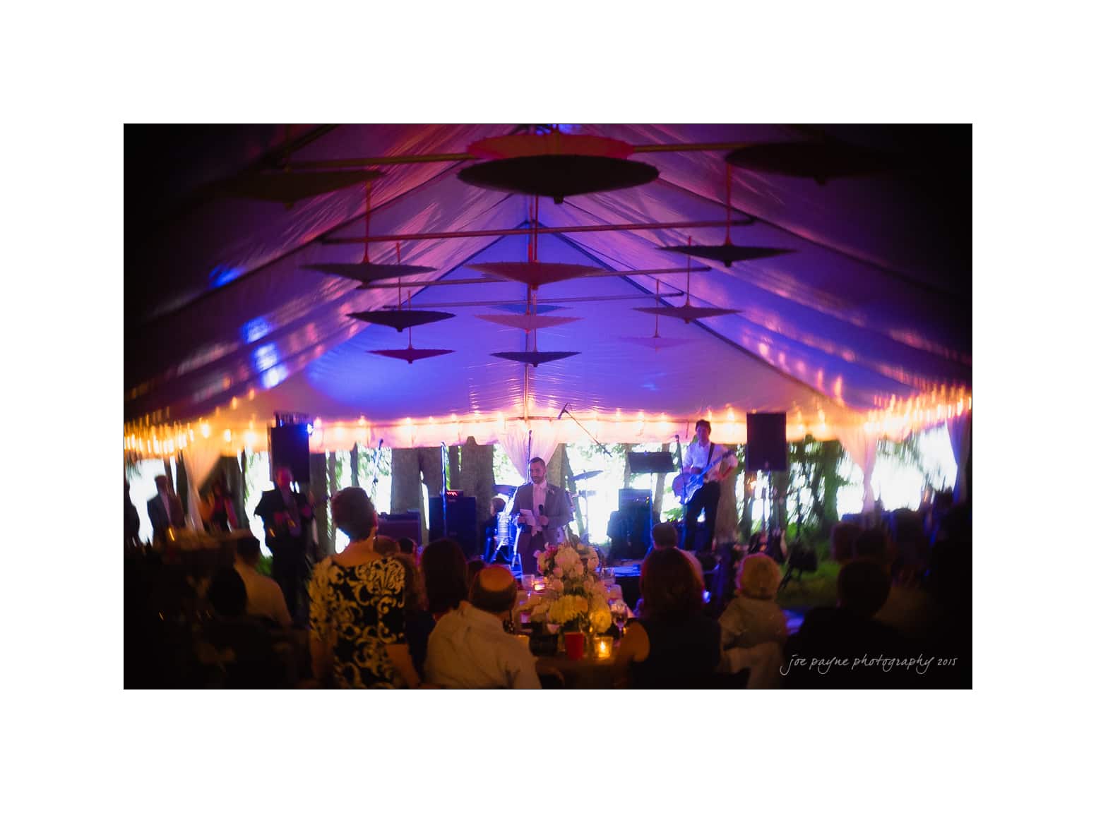 NC wedding photography - rachel & joe A band performing on stage under a large tent with colorful lights. The audience is seated at tables, watching the show. The atmosphere is lively and festive, with hanging decorations and a warm glow from string lights.