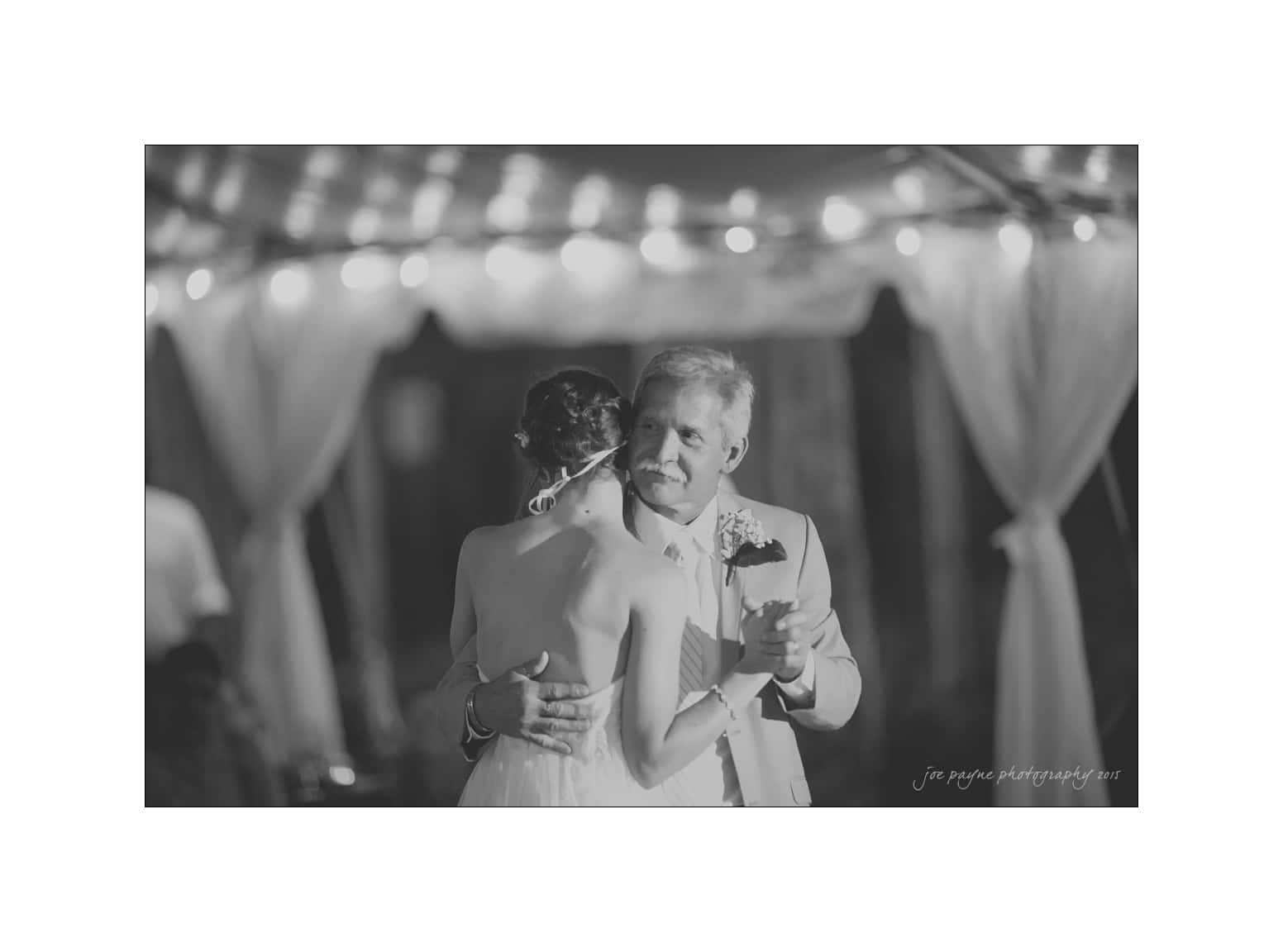NC wedding photography - rachel & joe A black and white photo of a bride and an older man embracing during a dance under string lights. The man holds a drink and a boutonniere is visible on his suit. Soft draped fabric is in the background, adding to the intimate atmosphere.