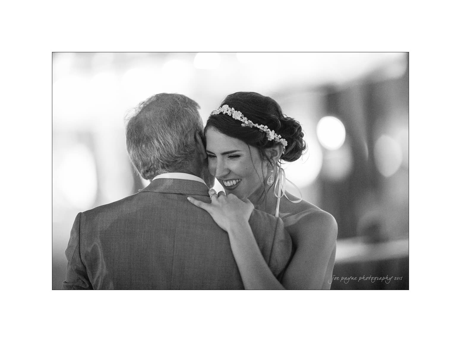 NC wedding photography - rachel & joe A black and white photo of a bride sharing a joyful moment dancing with an older man. She wears a floral headpiece and earrings, smiling with her eyes closed as they embrace gently.