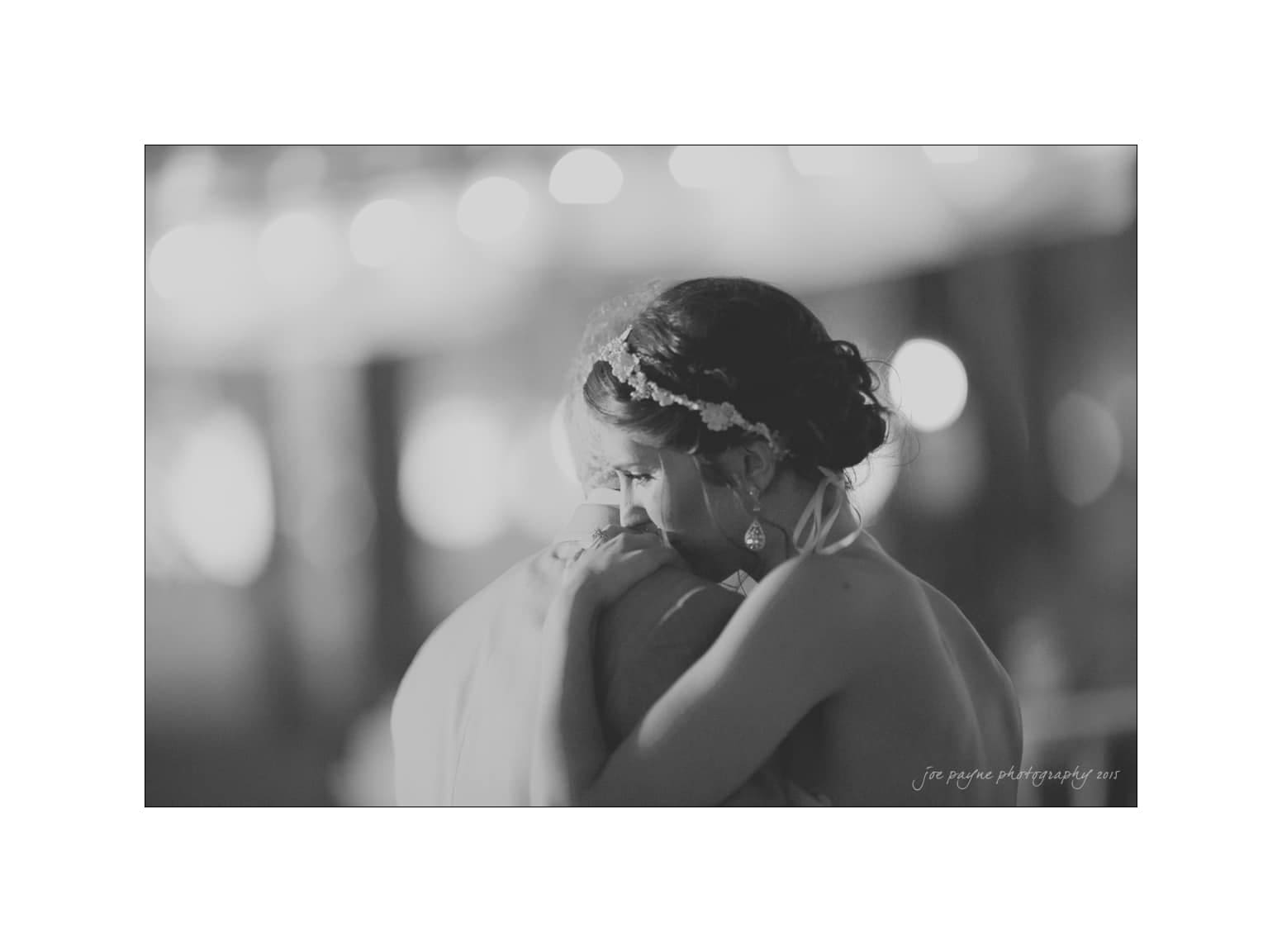 NC wedding photography - rachel & joe A black-and-white image showing a couple gently embracing, with the woman resting her head on the mans shoulder. She wears a hairpiece and earrings, creating an intimate and tender atmosphere. The background is softly blurred with bokeh lights.