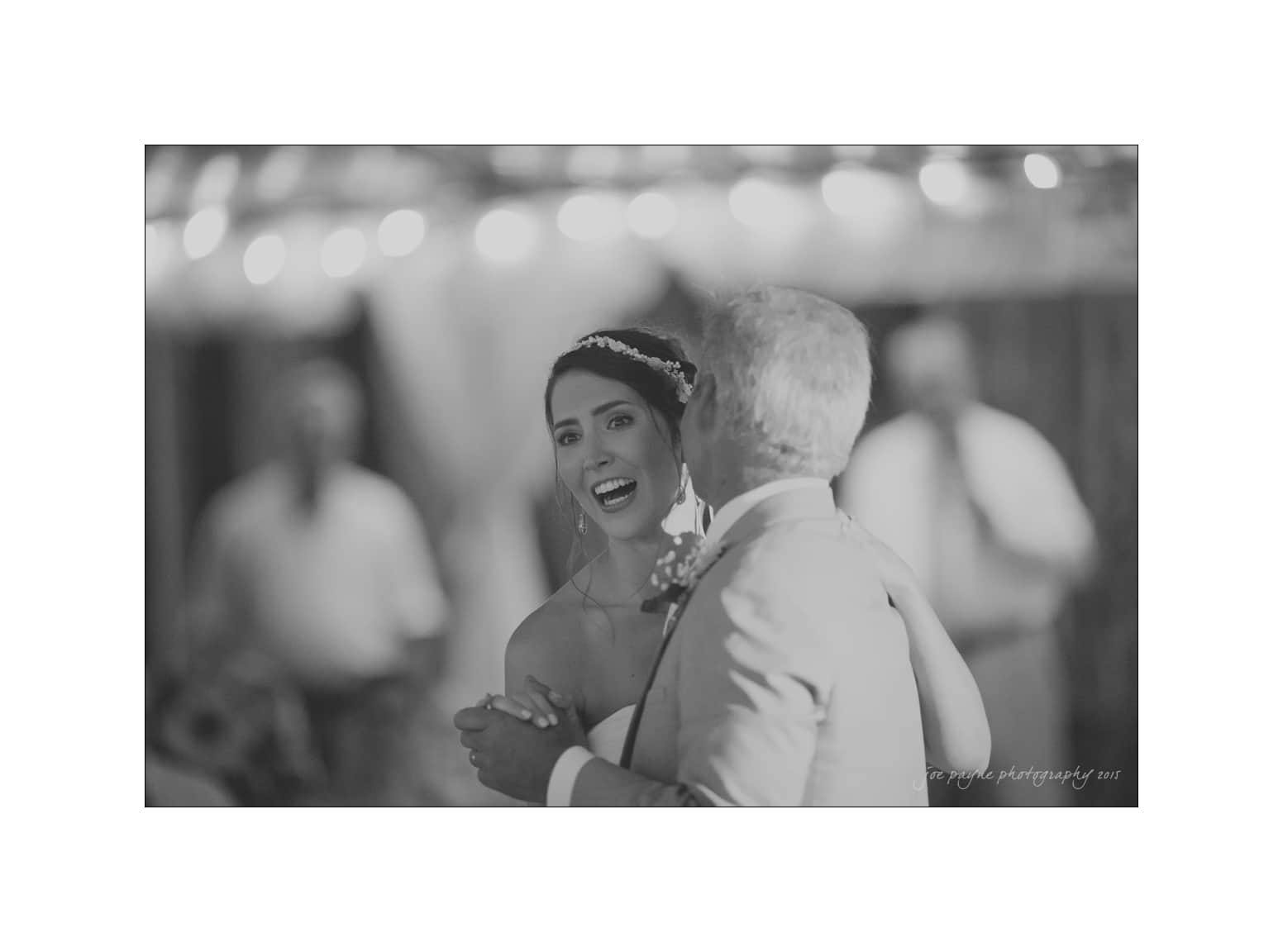 NC wedding photography - rachel & joe A bride joyfully dances with an older man in a black and white photo. String lights softly illuminate the background, creating a warm and celebratory atmosphere.