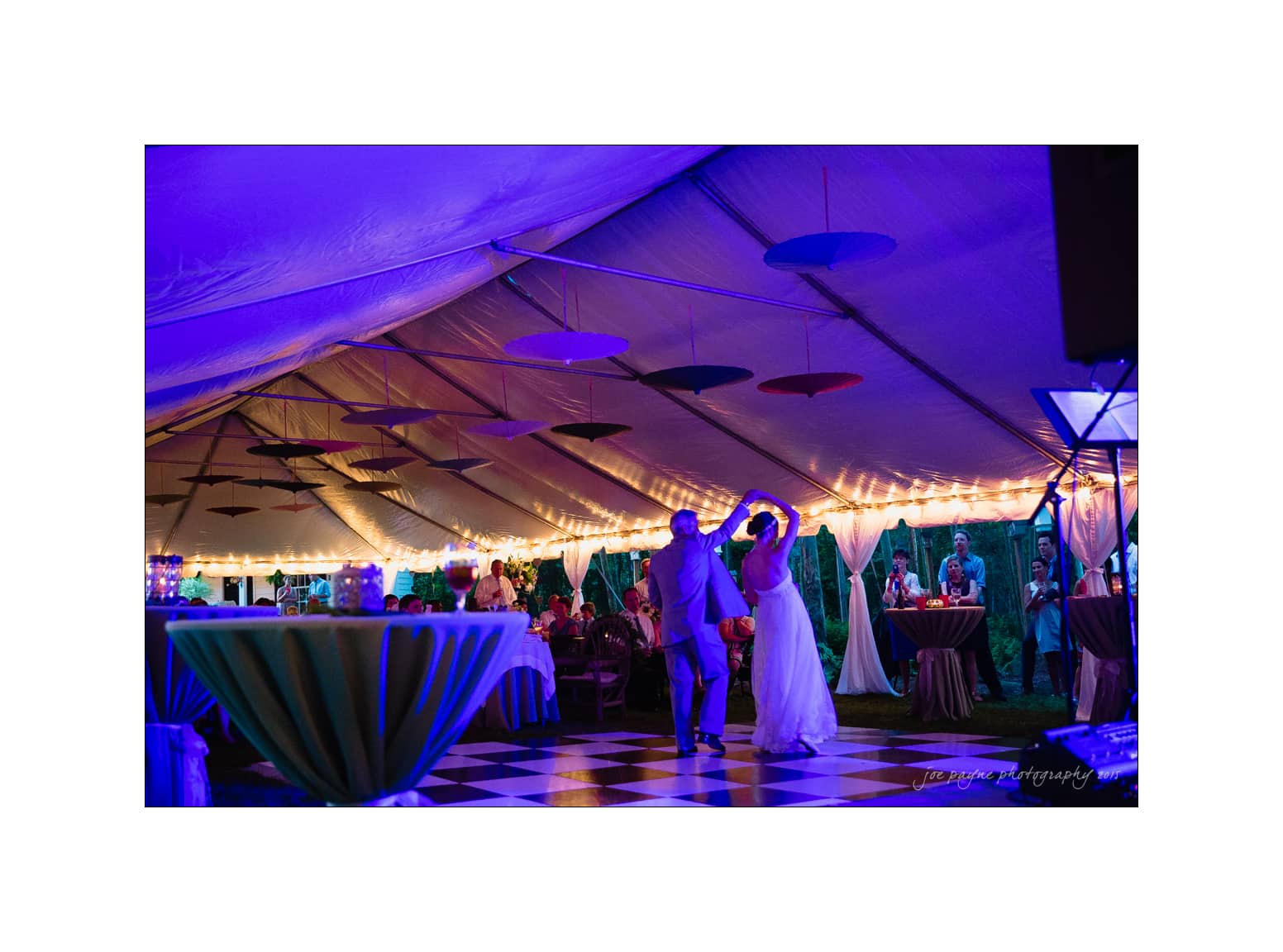 NC wedding photography - rachel & joe A couple dances under a large, illuminated tent at night. The tent is decorated with hanging umbrellas and string lights. A checkerboard dance floor and round tables are visible, with guests sitting and enjoying the event.