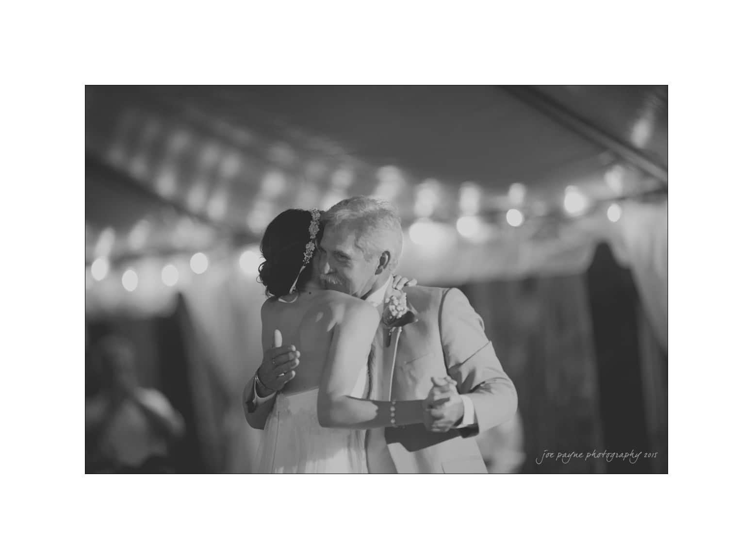 NC wedding photography - rachel & joe A groom lovingly embraces his bride during their first dance at a wedding reception. The scene is illuminated by soft, romantic lighting and string lights in the background, creating a warm and intimate atmosphere.
