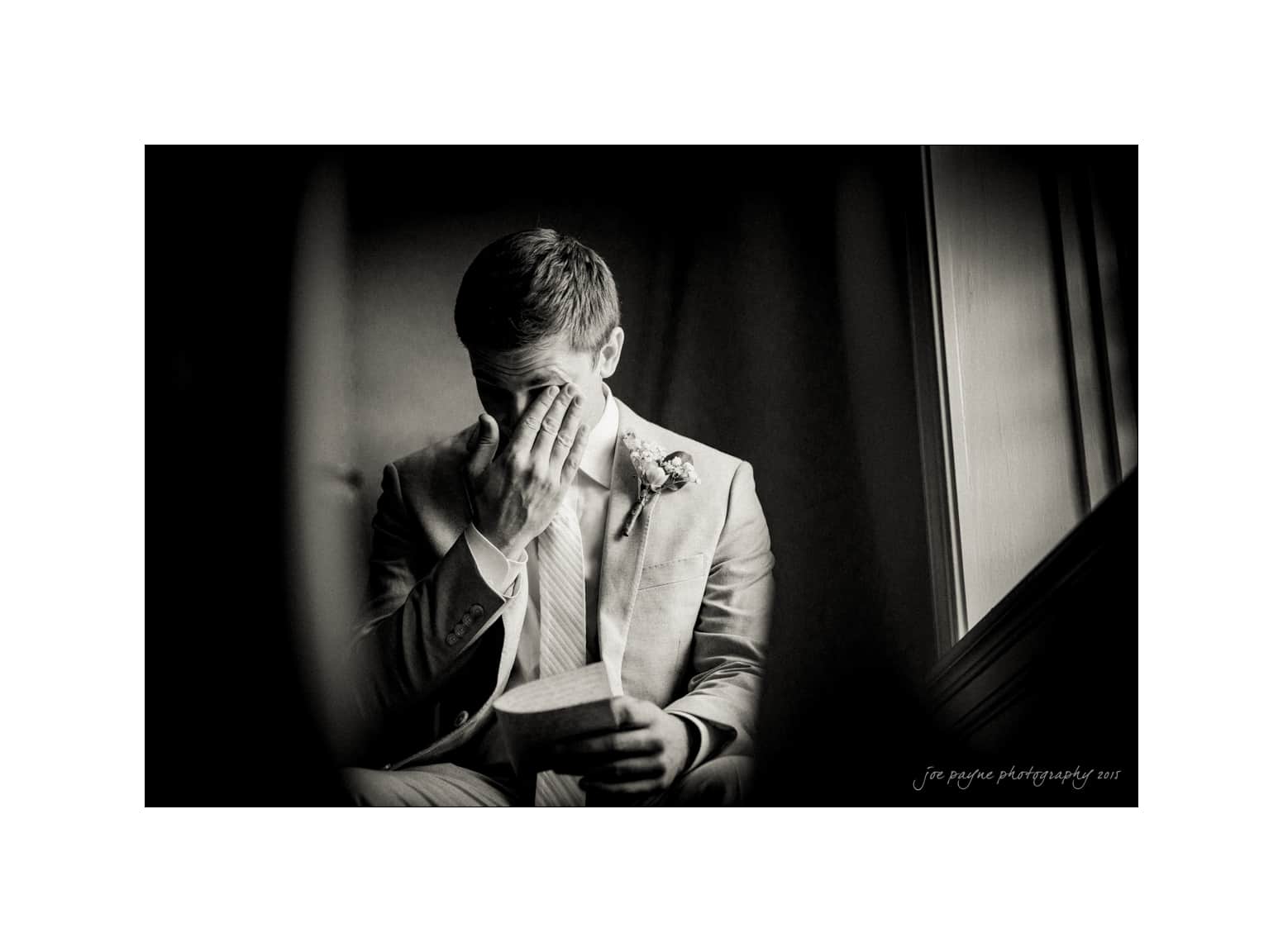 NC wedding photography - rachel & joe A black and white photo of a man in a suit sitting by a window, covering his face with one hand, holding a piece of paper in the other. It appears he is emotional or reflecting deeply. Theres a flower pinned to his lapel.