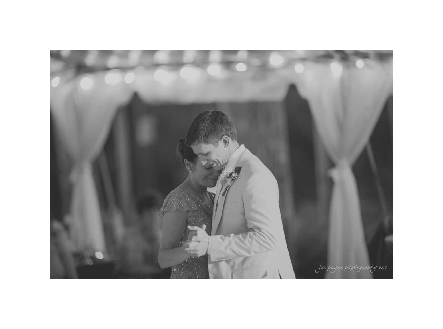 NC wedding photography - rachel & joe A black and white photo of a couple dancing closely under string lights. The man wears a suit, and the woman is in a dress with delicate details. They look content and share an intimate moment, surrounded by soft, draped fabric in the background.