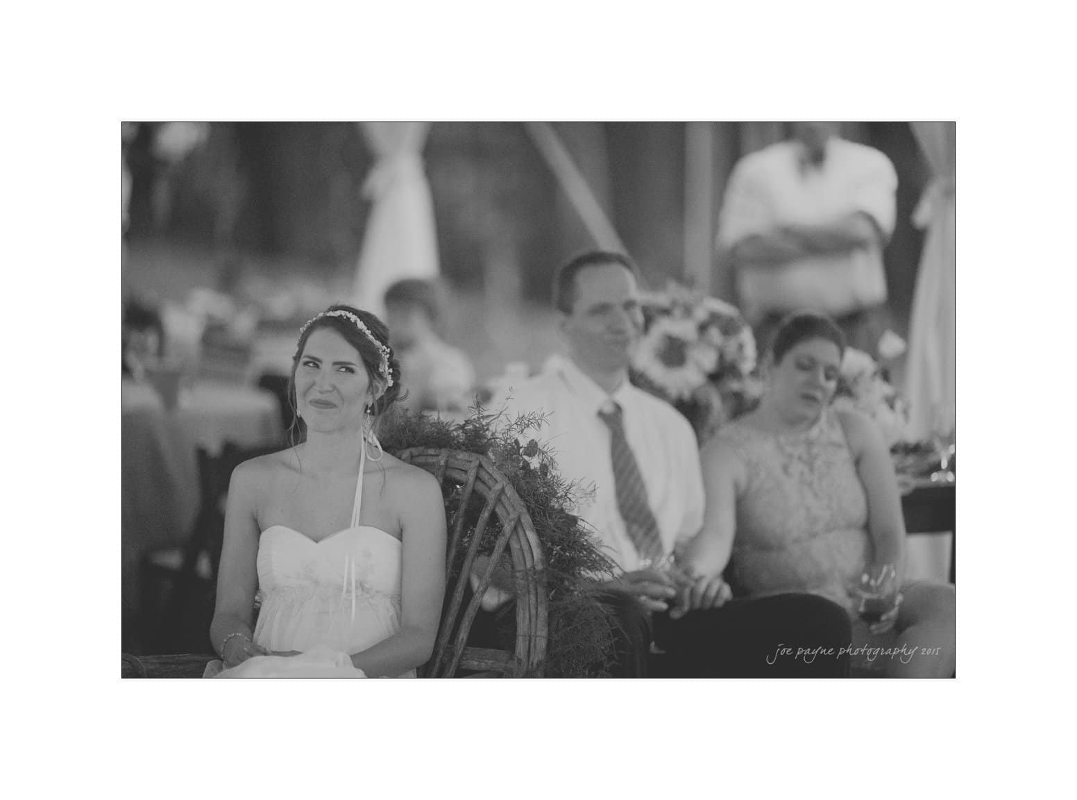 NC wedding photography - rachel & joe A bride in a white dress sits smiling on a wooden chair outdoors. Behind her, a man in a tie appears relaxed, and a woman in a lace dress rests her head on his shoulder. The setting is a casual outdoor event.