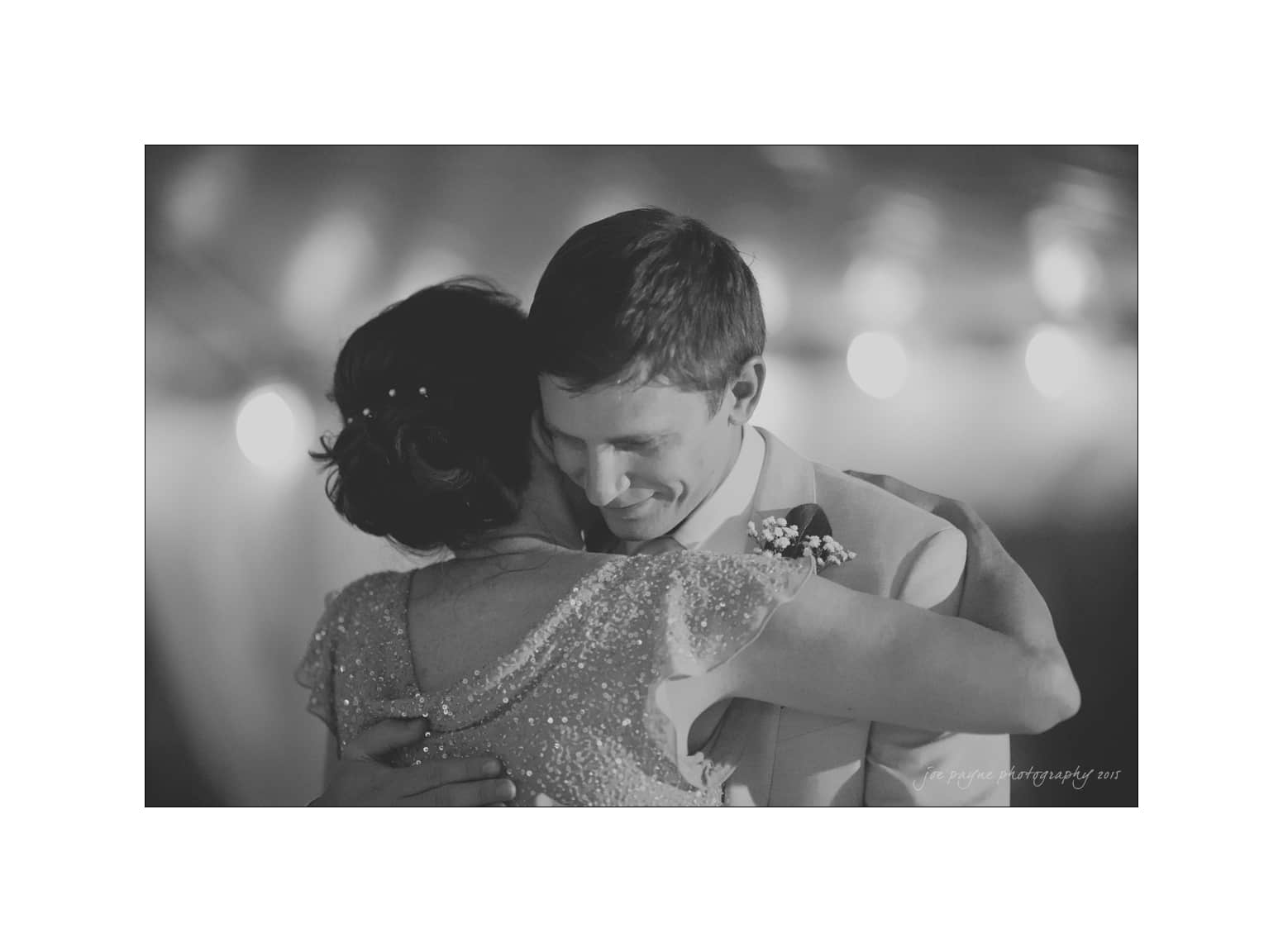 NC wedding photography - rachel & joe A black and white photo of a couple embracing during a dance. The man is wearing a suit with a boutonniere, and the woman is wearing a sparkly dress. The background is softly lit, creating an intimate atmosphere.