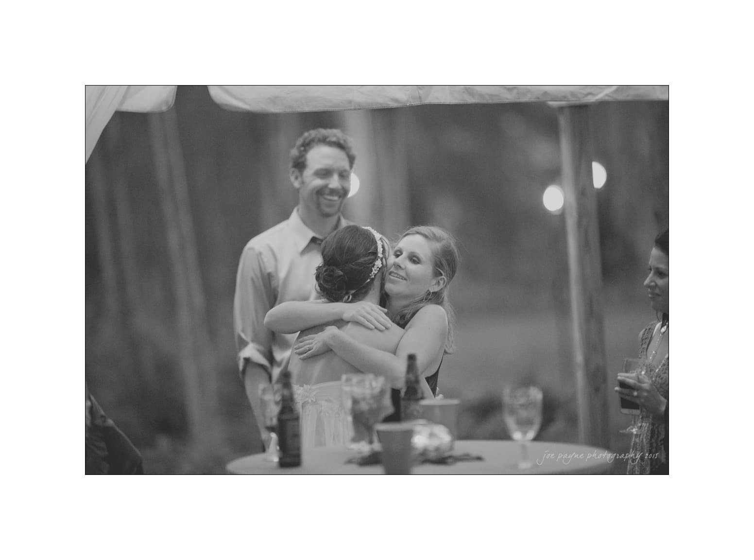 NC wedding photography - rachel & joe A black and white photo of two women hugging at an outdoor gathering, surrounded by a smiling man and another person at a table with drinks. The atmosphere appears joyful and celebratory.