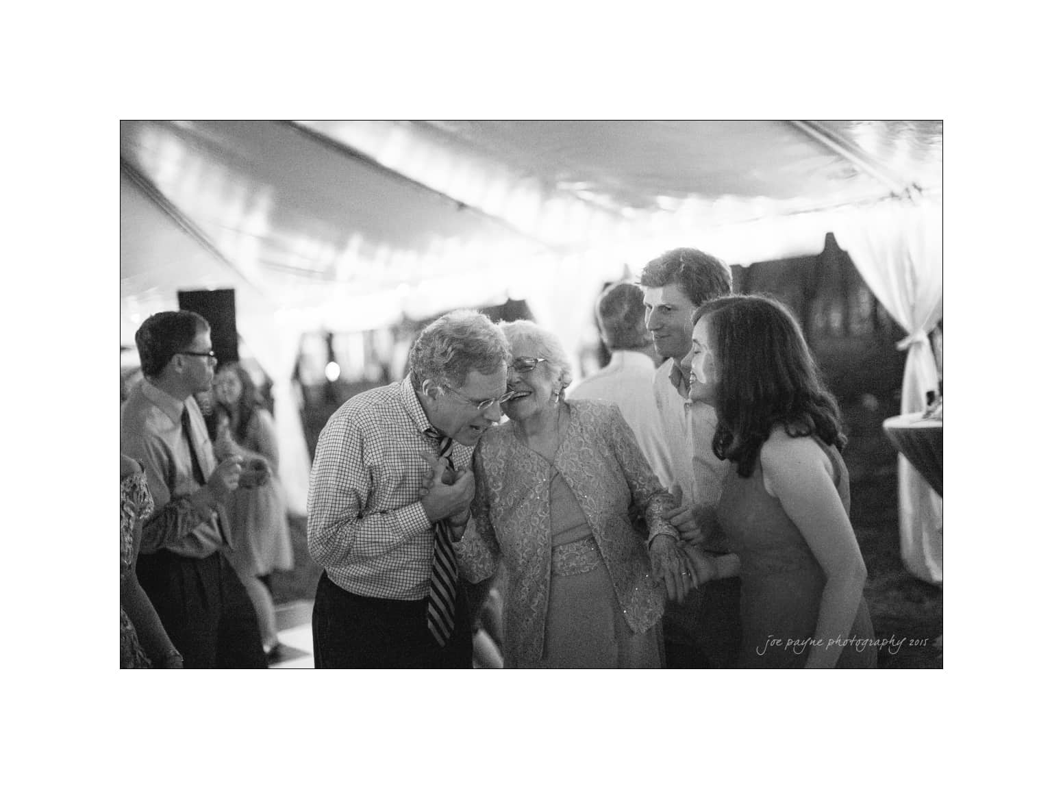 NC wedding photography - rachel & joe A group of people laughing and engaging under a tent at an event. The scene is in black and white, with festive lights in the background. The group appears to be enjoying a moment together.