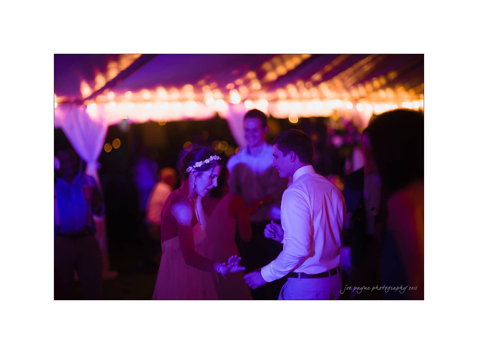 NC wedding photography - rachel & joe A couple dances under purple and orange string lights at an outdoor event. The woman wears a floral crown and a dress, while the man is in a white shirt. Other guests are blurred in the background, adding to the festive atmosphere.