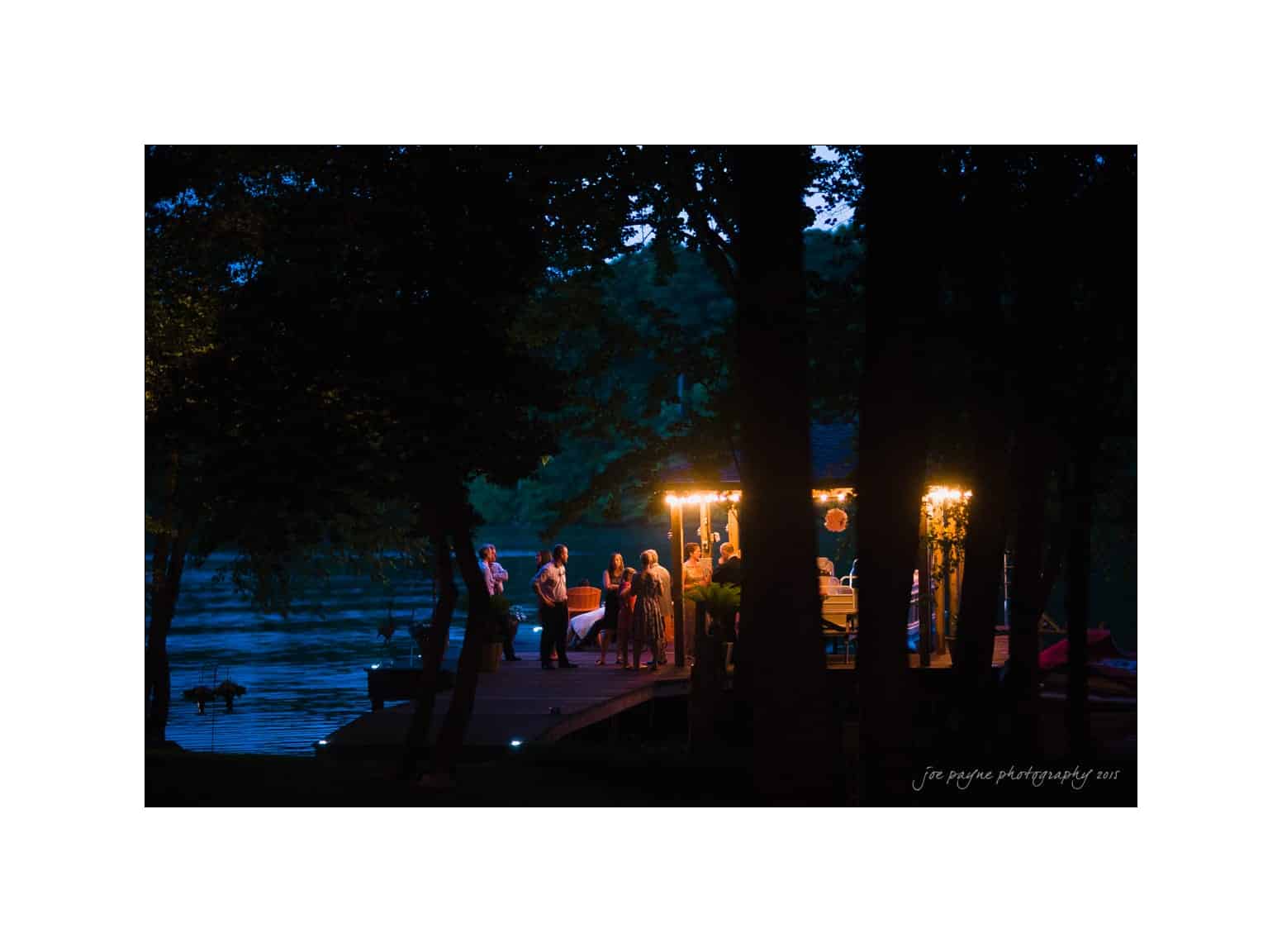 NC wedding photography - rachel & joe A small group of people gather on a dimly lit dock at night by a lake. Trees frame the scene, and soft lights from a canopy illuminate the group, creating a cozy ambiance against the serene water backdrop.