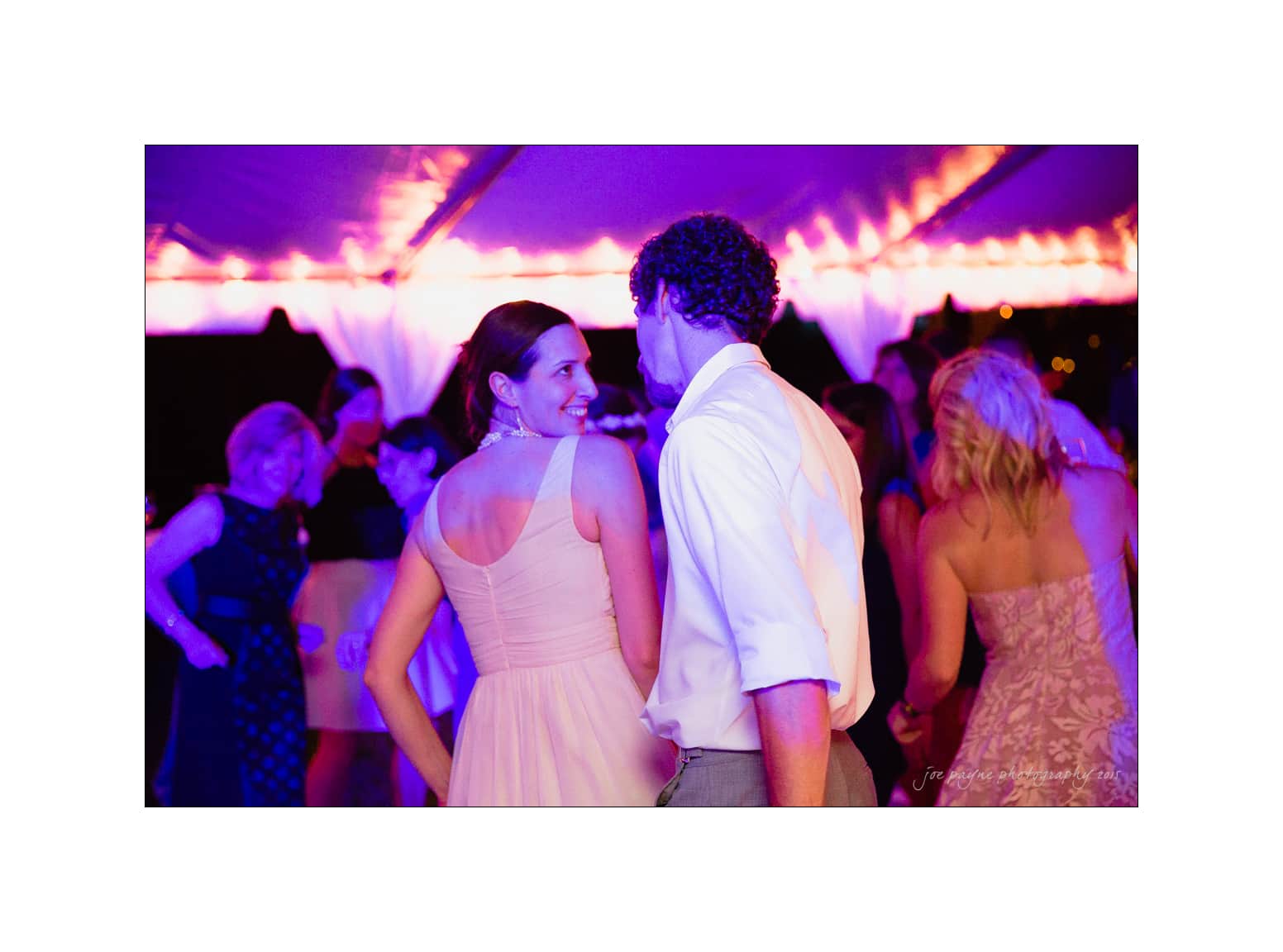 NC wedding photography - rachel & joe A couple dances closely under a canopy with purple lighting. The woman in a light pink dress and the man in a white shirt are smiling at each other. Other people are dancing in the background, creating a lively atmosphere.