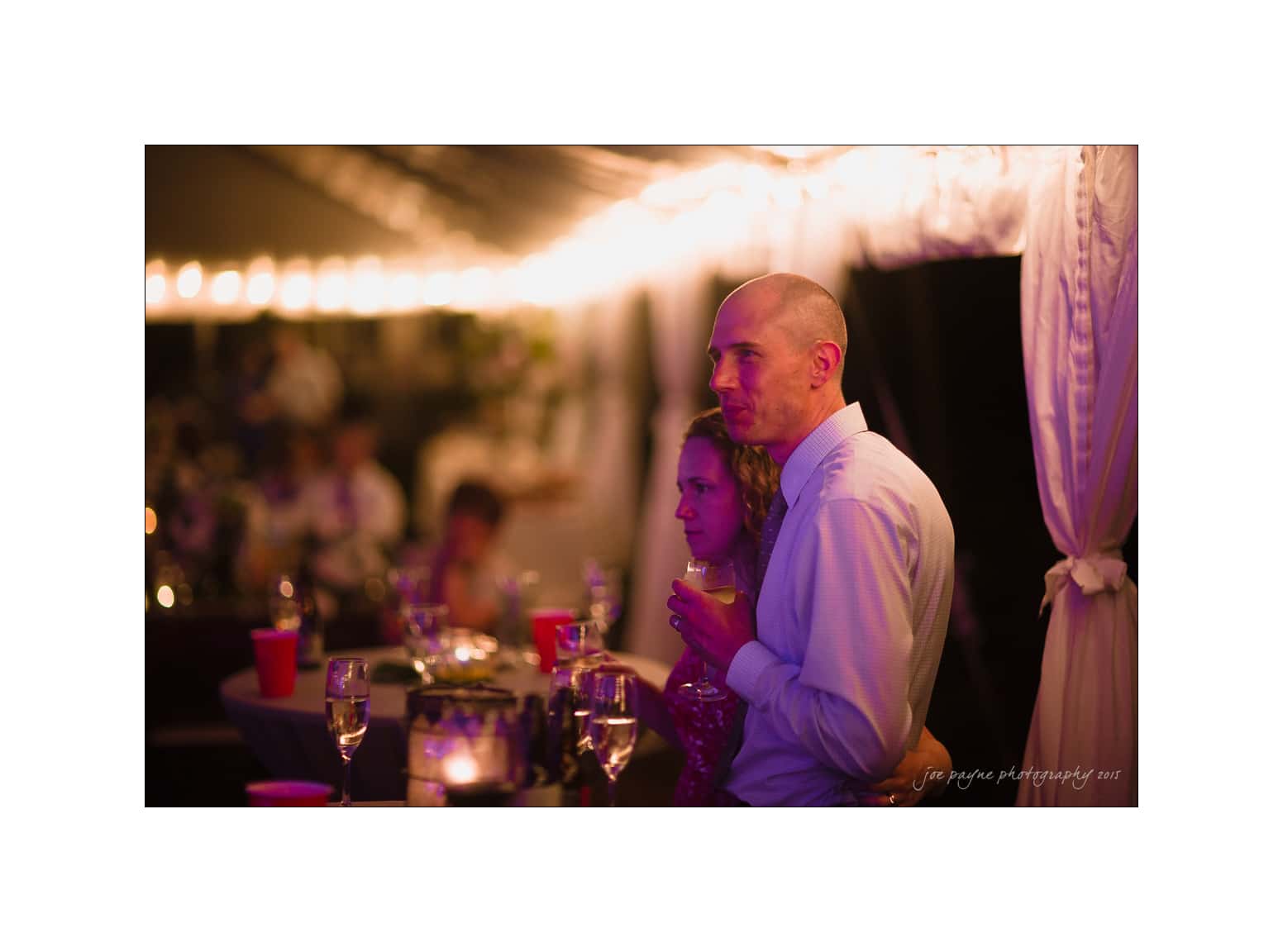 NC wedding photography - rachel & joe A couple stands closely together under warm, ambient lighting at a festive gathering. They hold drinks and gaze attentively at something out of the frame. A table with candles and red cups is nearby, and the background is softly blurred.