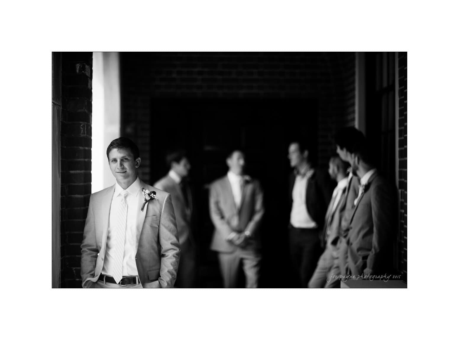 NC wedding photography - rachel & joe A groom in a suit stands in focus on the left, smiling, with a flower boutonniere. In the background, four groomsmen in similar suits stand and talk, slightly blurred. The setting appears to be indoors with a brick wall.
