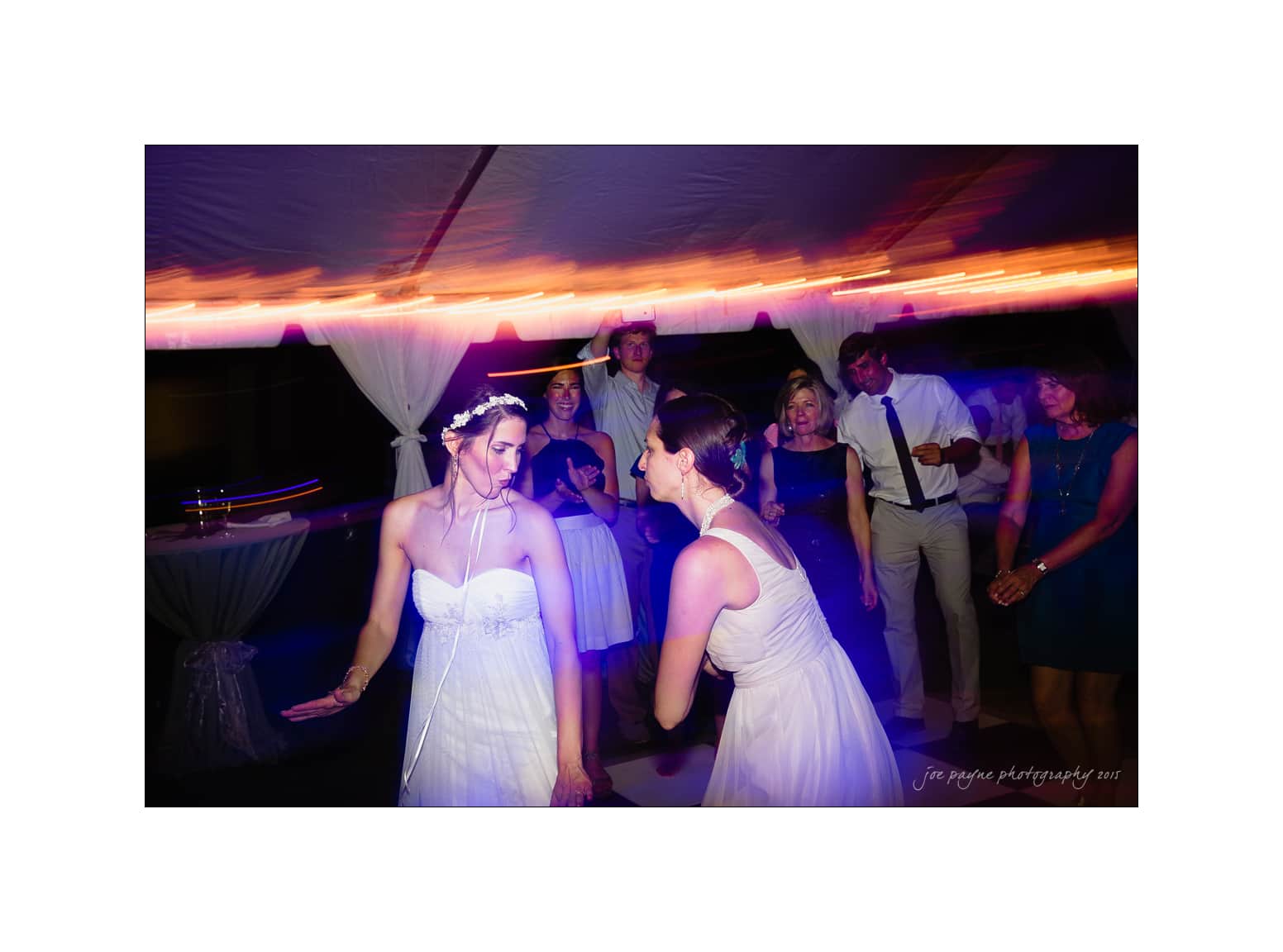 NC wedding photography - rachel & joe Two brides dance joyfully at their wedding reception under a tent. Guests surround them, watching and smiling. The scene is lively, with colorful lights creating a festive atmosphere.