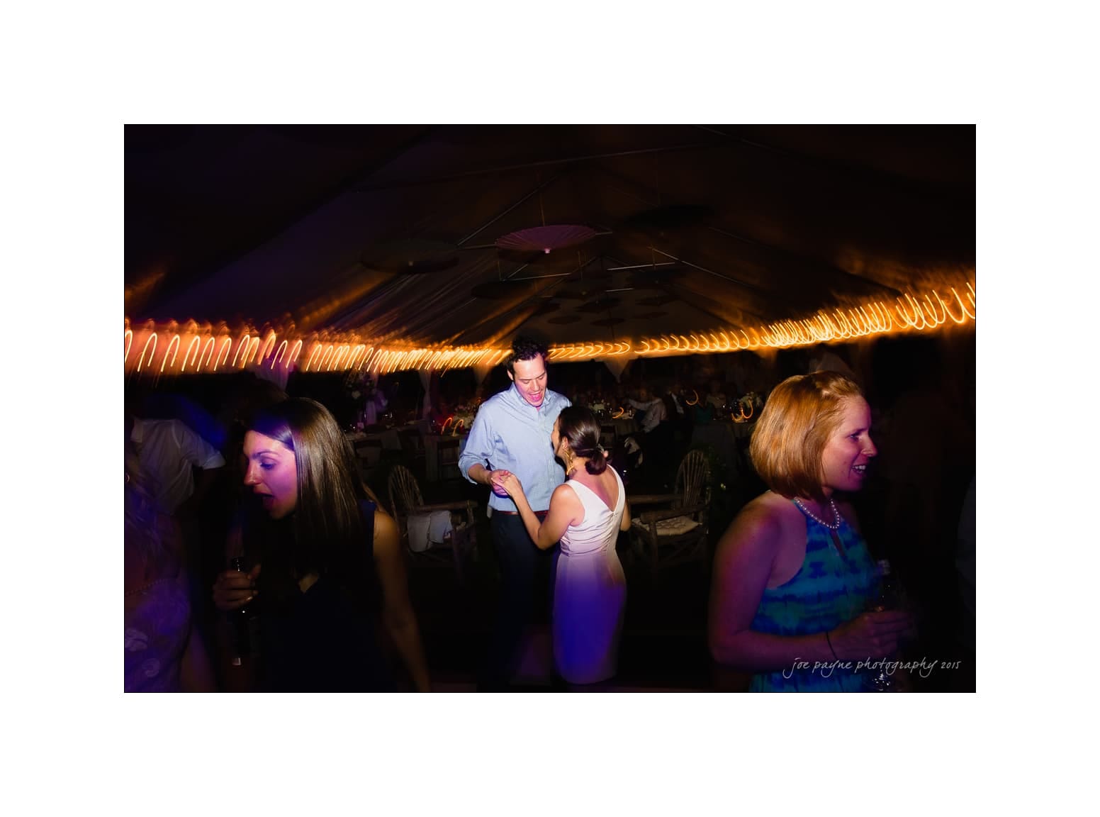 NC wedding photography - rachel & joe A lively party scene with people dancing under warm string lights. The room is dimly lit, creating a vibrant atmosphere. One couple dances in the center, while others socialize around them. People are dressed in semi-formal attire.