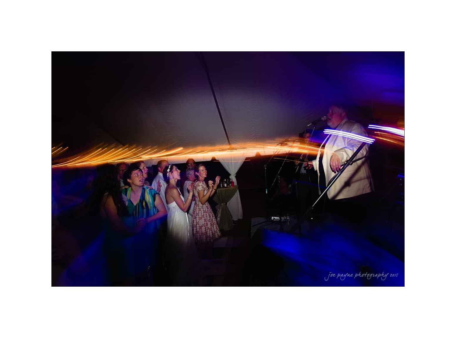 NC wedding photography - rachel & joe A lively scene at a wedding reception with a bride and guests dancing near a stage. The band performs under a tent with vibrant blue and orange lights, creating a festive and dynamic atmosphere.