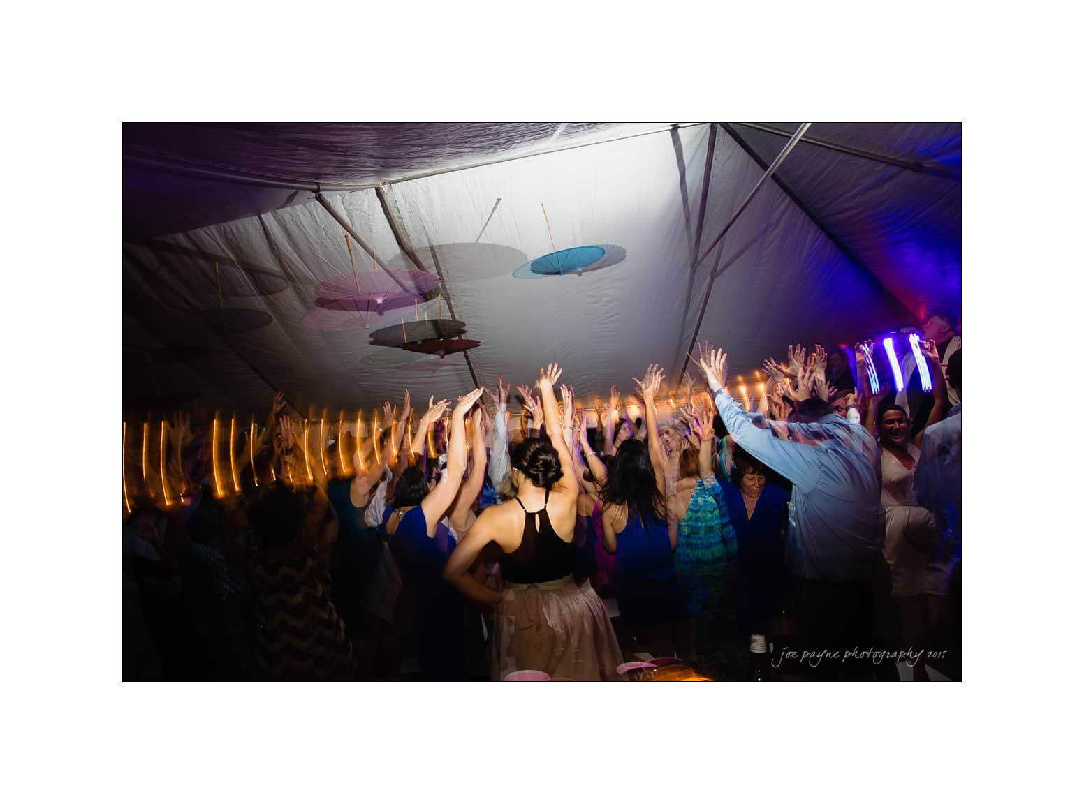 NC wedding photography - rachel & joe A lively crowd of people raise their hands and dance under a large illuminated tent, with colorful paper lanterns hanging from the ceiling. The scene is vibrant and filled with joyful energy.