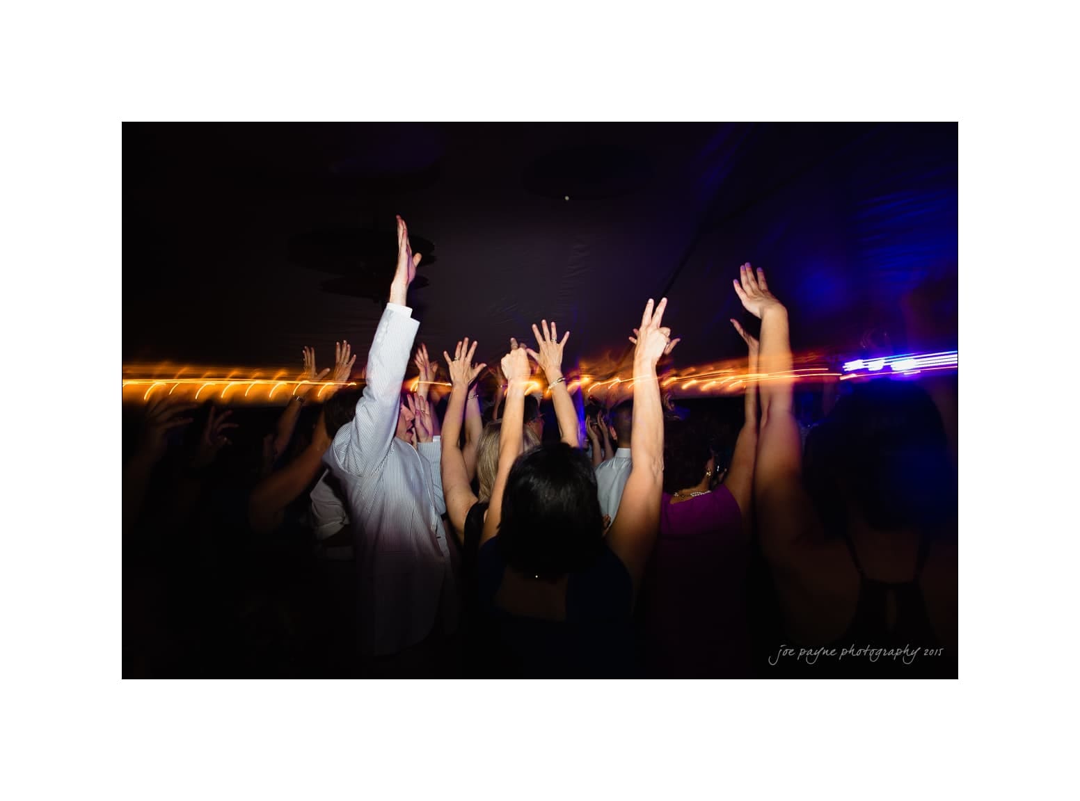 NC wedding photography - rachel & joe A group of people with raised hands dance energetically under colorful lights at a party or concert. The image captures motion with light streaks and lively ambiance.
