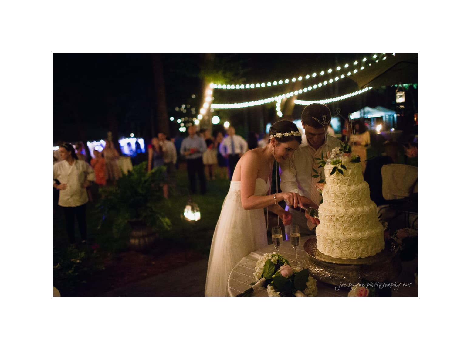 NC wedding photography - rachel & joe A bride and groom cut their wedding cake under string lights at an outdoor evening reception. Guests are blurred in the background, with warm lighting creating a cozy atmosphere.