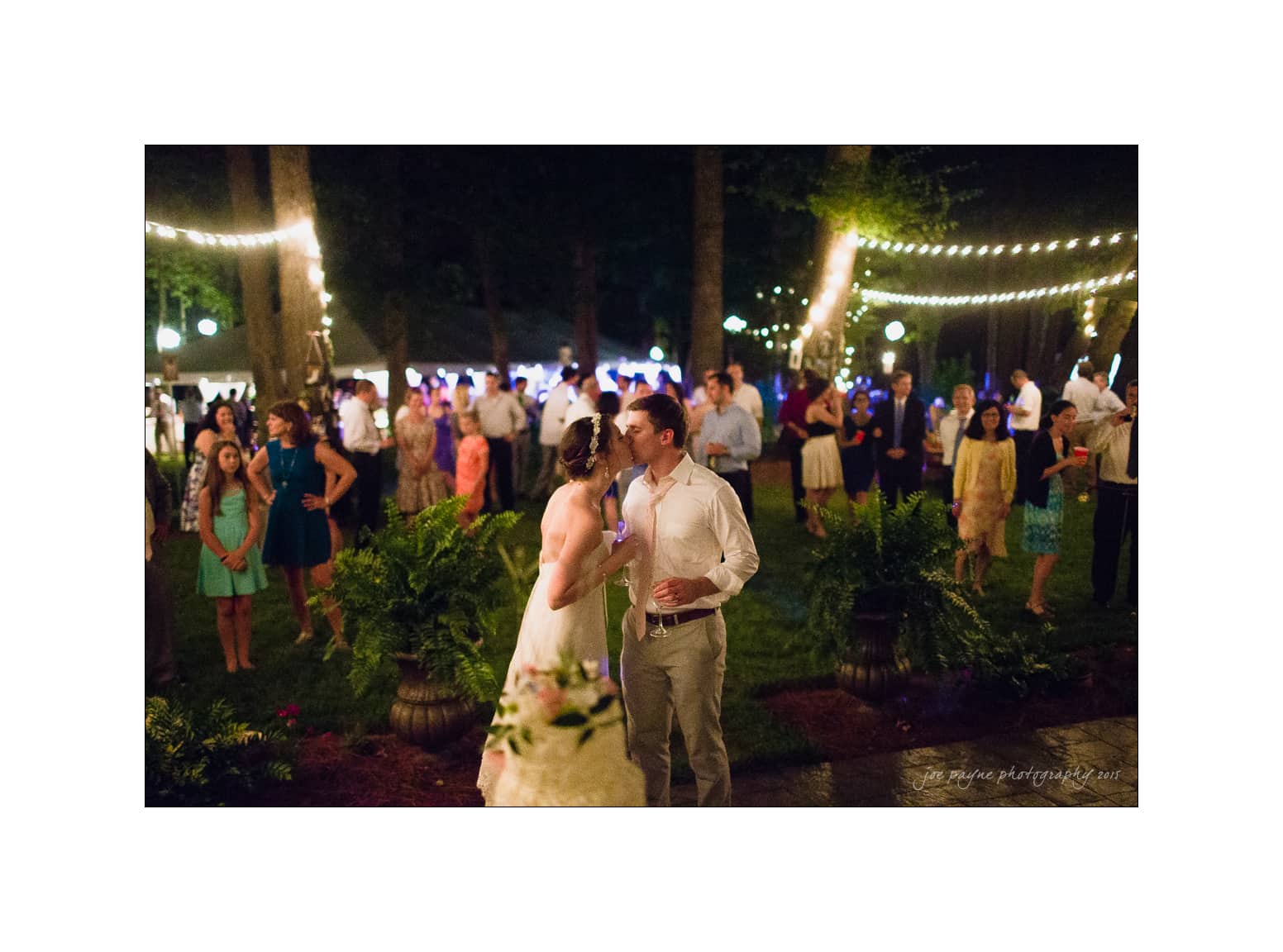 NC wedding photography - rachel & joe A bride and groom share a kiss at an outdoor night wedding reception. They stand under string lights, surrounded by guests who watch them. The scene is festive, with a warm, intimate atmosphere.