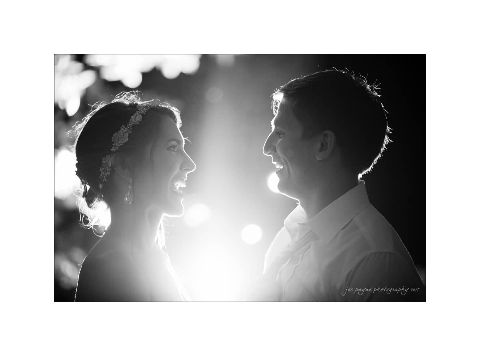 NC wedding photography - rachel & joe A black and white photo of a couple facing each other, smiling warmly, with soft backlighting creating a radiant glow around them. The woman wears a headband and the man is in a shirt, both exuding joy and affection.