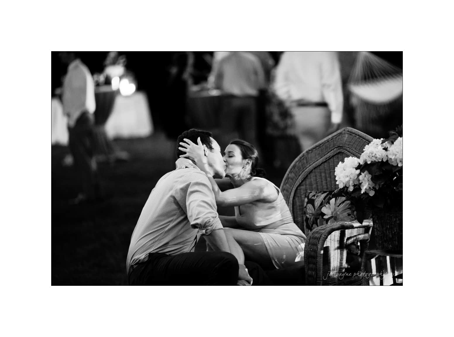 NC wedding photography - rachel & joe A black and white photo of a couple sitting closely. The woman leans in to kiss the man while holding his face. They are outdoors with blurred figures and a hammock in the background. A wicker chair with flowers is to their side.