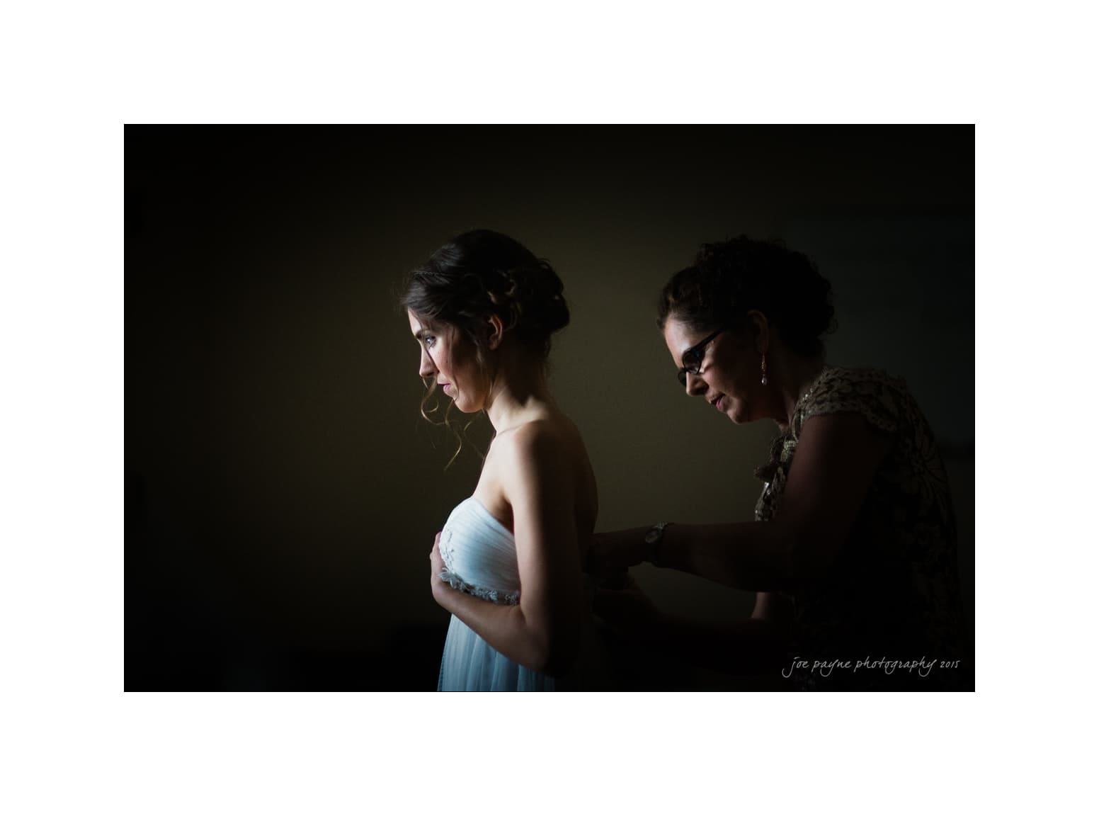 NC wedding photography - rachel & joe A woman in a light dress stands in a dimly lit room as another woman, wearing glasses, helps fasten the back of her dress. The mood is calm and focused, with a soft light illuminating their profiles.