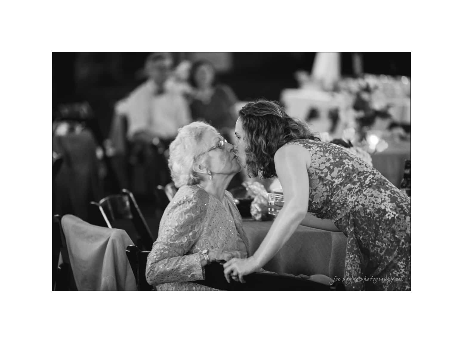 NC wedding photography - rachel & joe A touching black-and-white photo of a young woman leaning in to kiss an elderly woman on the cheek at a gathering. Both are smiling, surrounded by dimly lit tables and guests in the background.