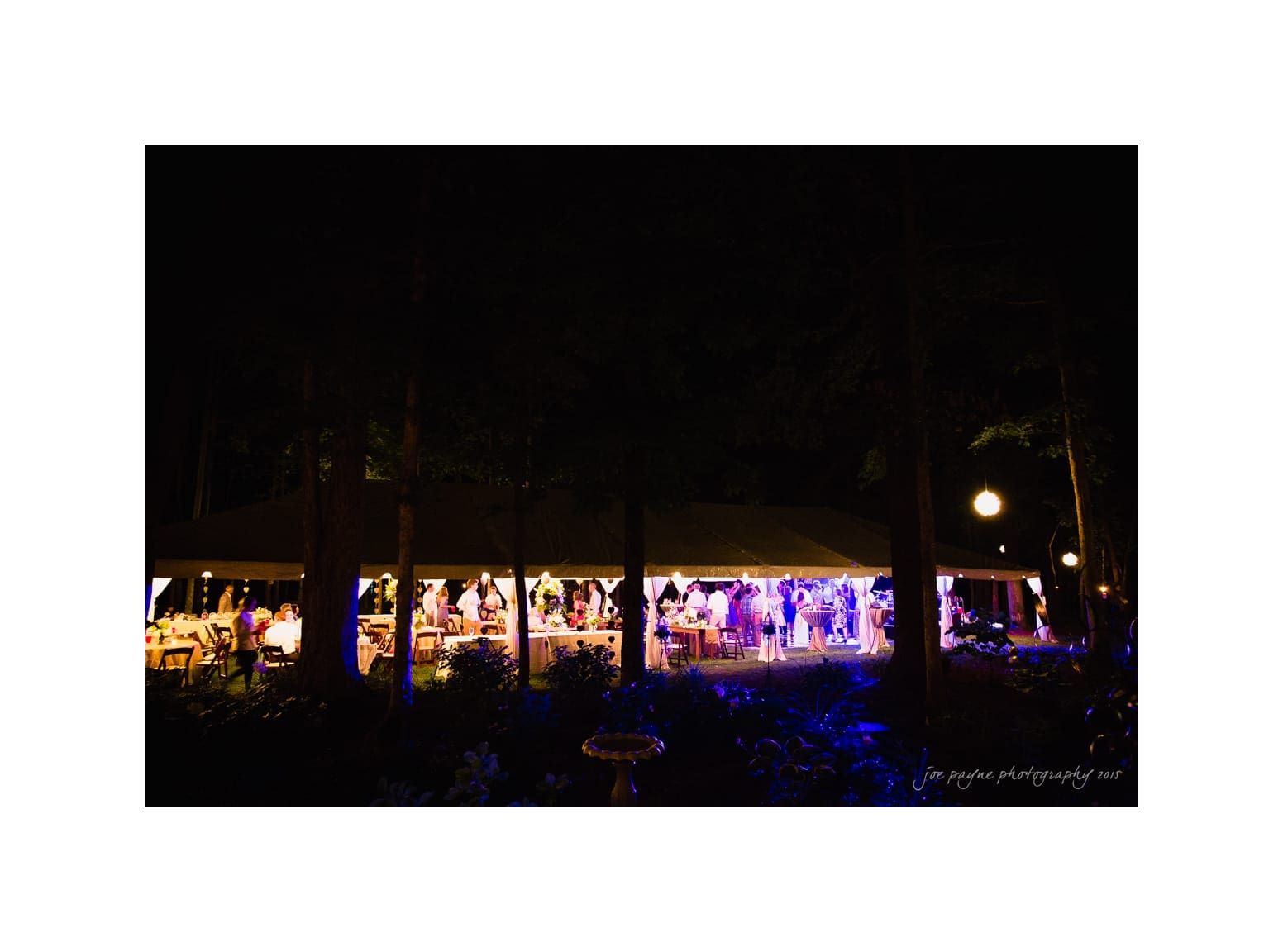 NC wedding photography - rachel & joe A nighttime scene of an outdoor event under a lit pavilion, surrounded by trees. The interior is illuminated with colorful lights, and groups of people are visible inside, engaging in various activities.