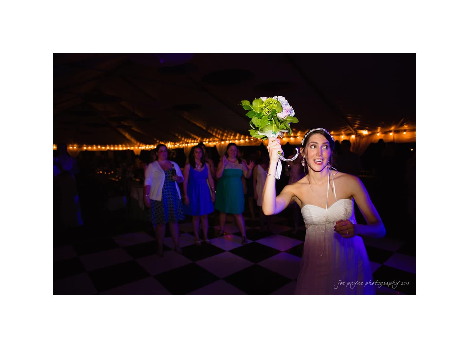 NC wedding photography - rachel & joe A bride in a white dress joyfully prepares to toss her bouquet over her shoulder at a wedding reception. Guests eagerly watch in the background, ready to catch it. The event is set in a warmly lit room with a checkered floor.
