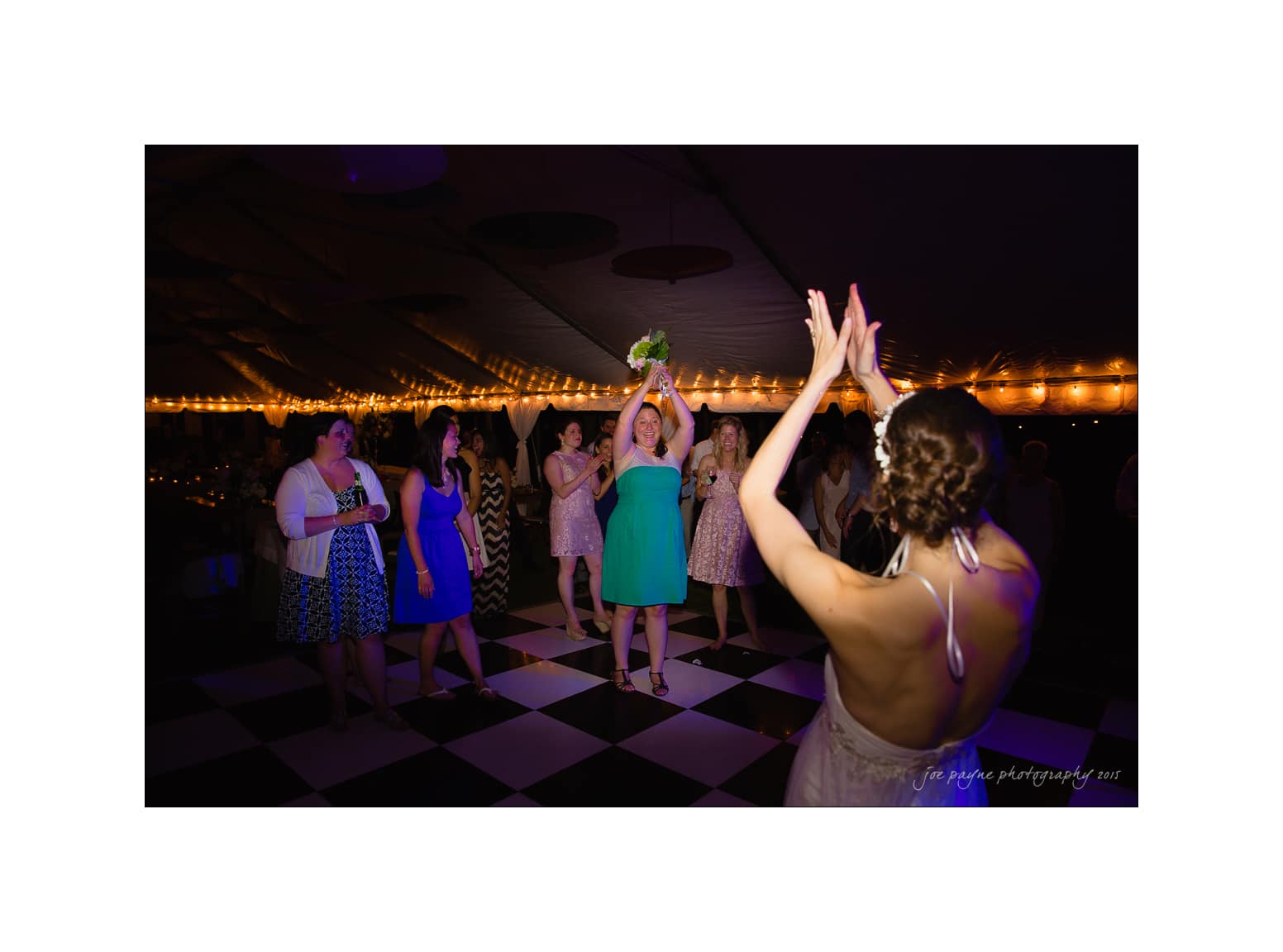 NC wedding photography - rachel & joe A bride tosses her bouquet on a checkered dance floor, surrounded by women eagerly reaching to catch it. The setting is a warmly lit tent with a festive atmosphere.