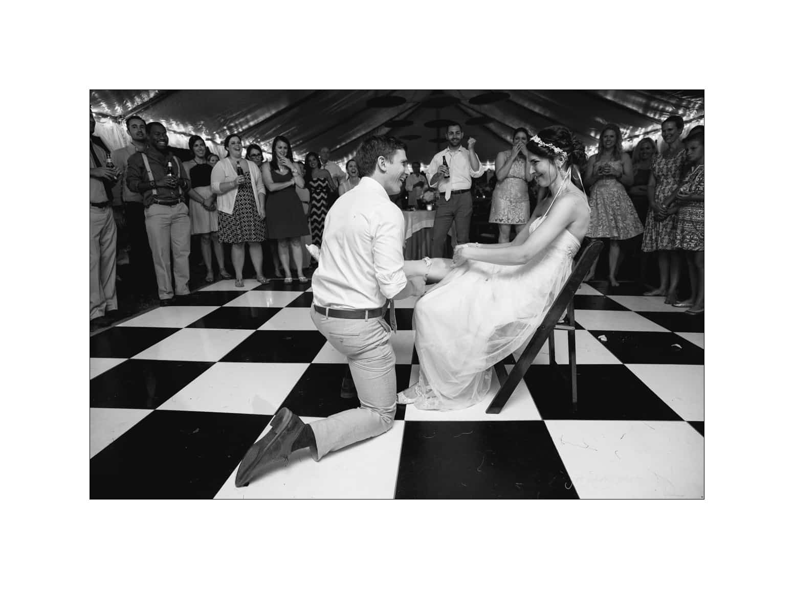 NC wedding photography - rachel & joe A groom kneels before the bride, seated on a chair, on a checkered dance floor at a wedding. Guests are gathered around, smiling and watching the interaction, under a tent illuminated with lights. The image is in black and white.