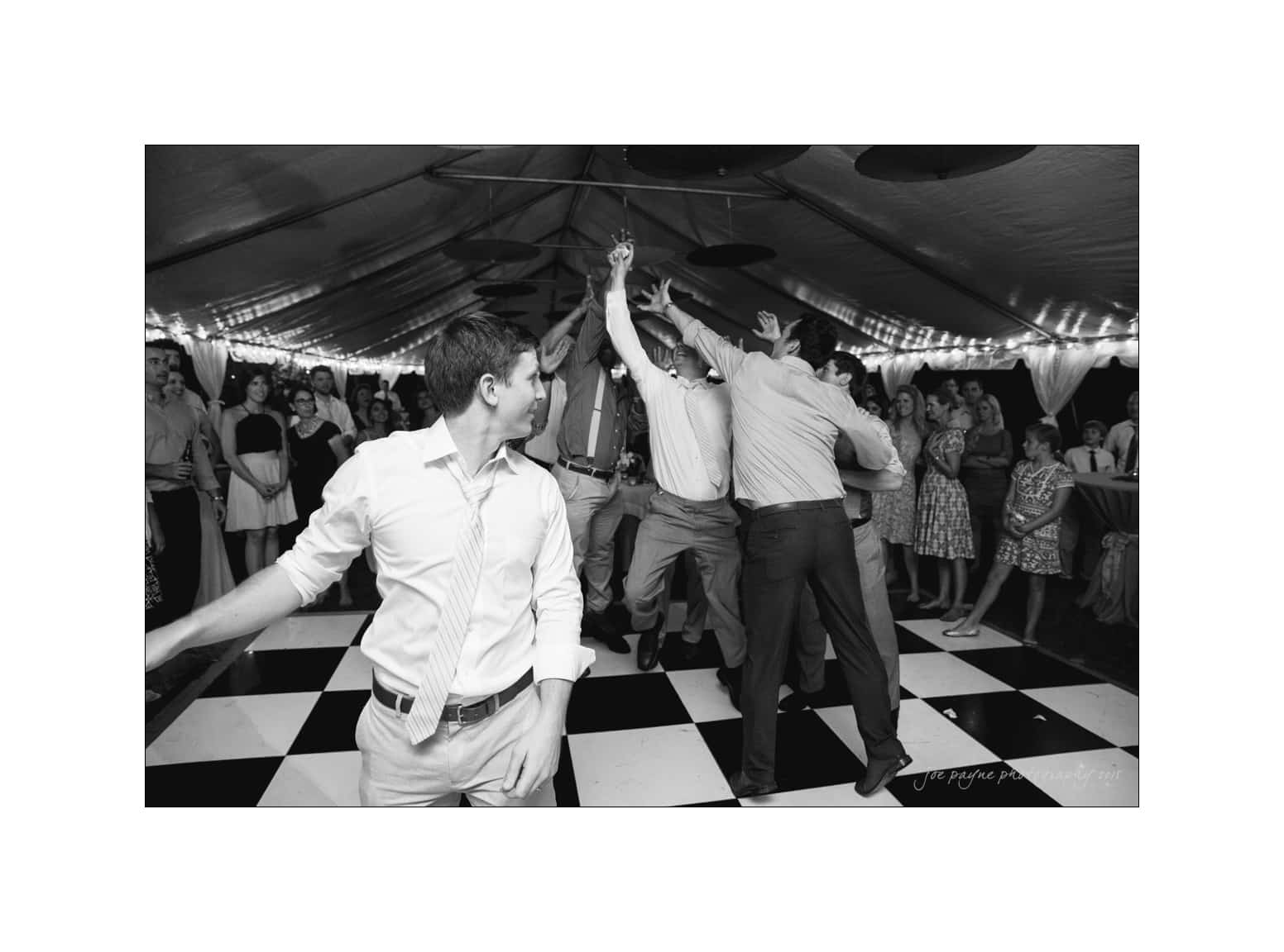 NC wedding photography - rachel & joe A black-and-white photo of a lively dance floor at a wedding reception. A group of people in formal attire enthusiastically reach towards the ceiling, surrounded by guests watching and smiling, under a tent decorated with lights.