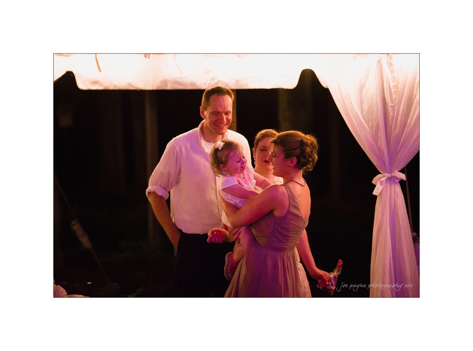 NC wedding photography - rachel & joe A joyful group at a nighttime outdoor event. A woman in a dress holds a smiling child, surrounded by two men and a girl. A tent canopy with soft lighting creates a warm ambiance.